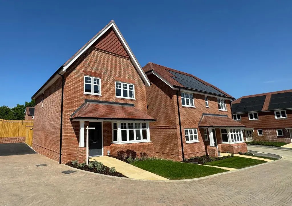 Row of modern brick houses beneath a clear blue sky. The homes feature white-framed windows, black front doors, and pitched roofs. The house on the left includes a garage with a paved driveway, while the house on the right displays solar panels on its roof. Both properties are fronted by neat lawns and simple landscaping along a quiet residential street.