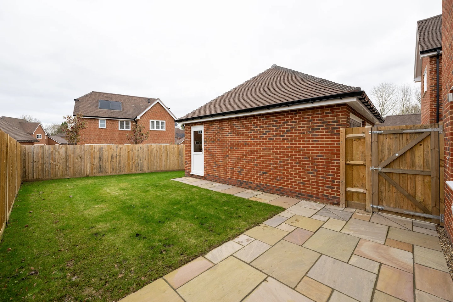 Enclosed backyard featuring a small rectangular lawn bordered by a paved patio. A wooden fence surrounds the space. At the rear, a brick outbuilding with a tiled roof includes a white door and a circular window. Neighboring houses are partially visible beyond the fence under an overcast sky.