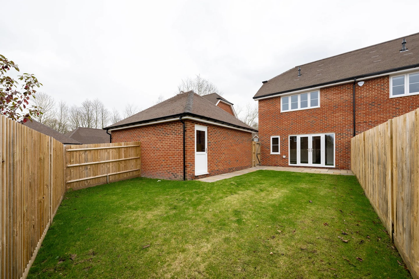 A neatly maintained suburban backyard enclosed by wooden fencing, featuring a well-kept green lawn. On the left, a small brick outbuilding with a circular window and a white door. To the right, a larger brick house with glass patio doors and several windows. The sky is overcast, and trees are visible beyond the fence.