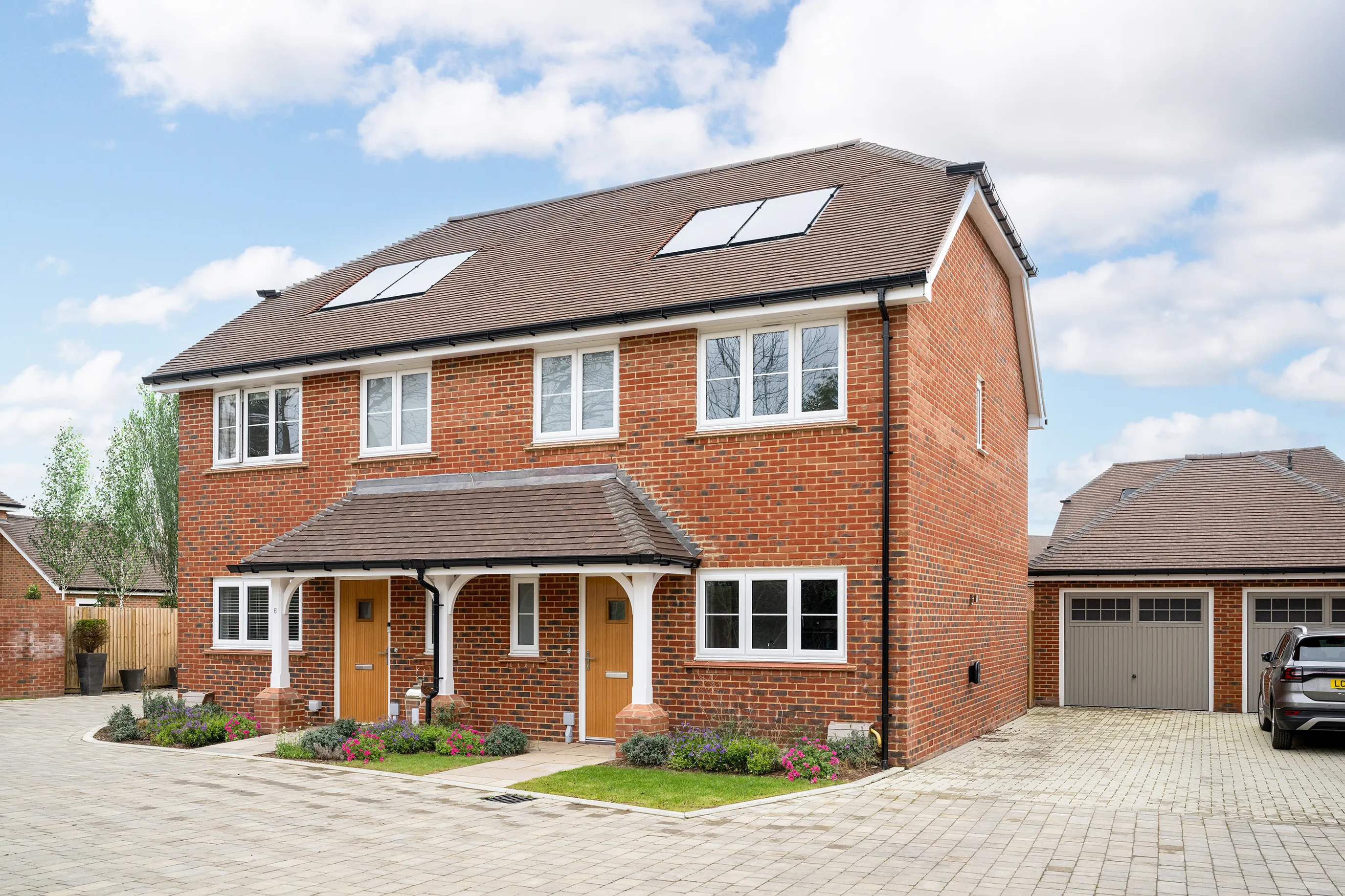 A semi-detached brick house with a pitched roof and solar panels on both sides. Two front doors indicate separate living units. The ground and first floors feature multiple white-trimmed windows. In front, a landscaped garden with flowers and bushes borders a paved driveway leading to a garage, where a silver car is parked. The sky is partly cloudy, and neighbouring houses appear in the background.