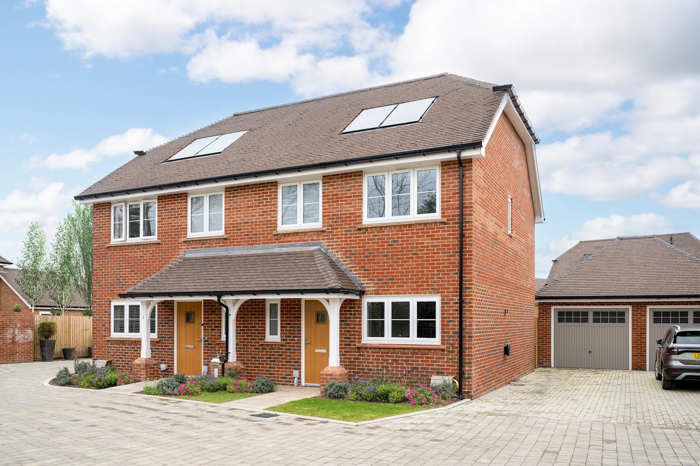 A semi-detached brick house with a pitched roof and solar panels on both sides. Two front doors indicate separate living units. The ground and first floors feature multiple white-trimmed windows. In front, a landscaped garden with flowers and bushes borders a paved driveway leading to a garage, where a silver car is parked. The sky is partly cloudy, and neighbouring houses appear in the background.
