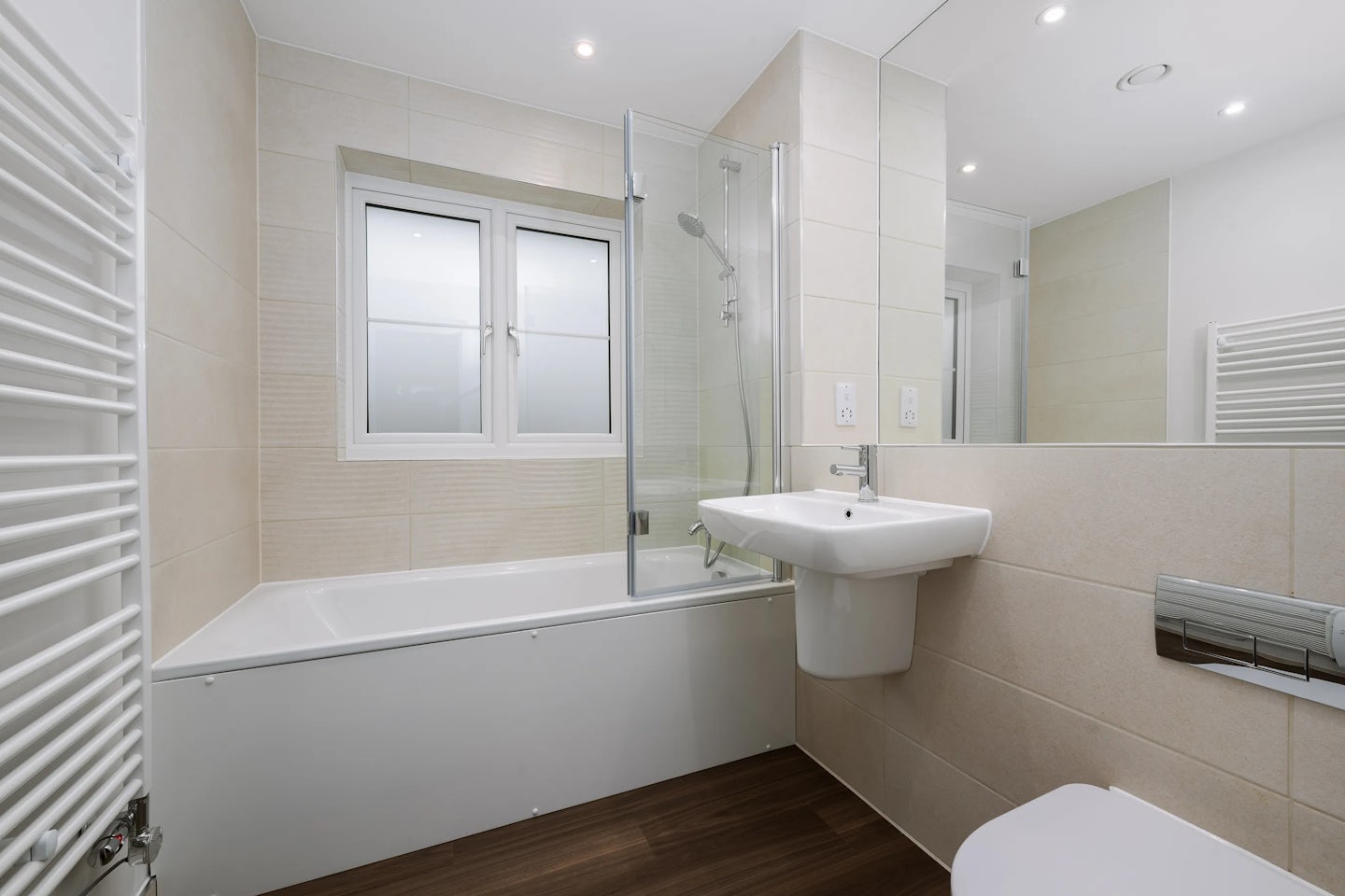 A modern, well-lit bathroom featuring a bathtub with a glass shower screen and wall-mounted showerhead beneath a window. The space includes a wall-mounted sink, a toilet, and a large mirror above the sink. The walls are tiled in light tones, while the flooring resembles dark wood. A white radiator is visible to the left, and recessed ceiling lights provide even illumination.