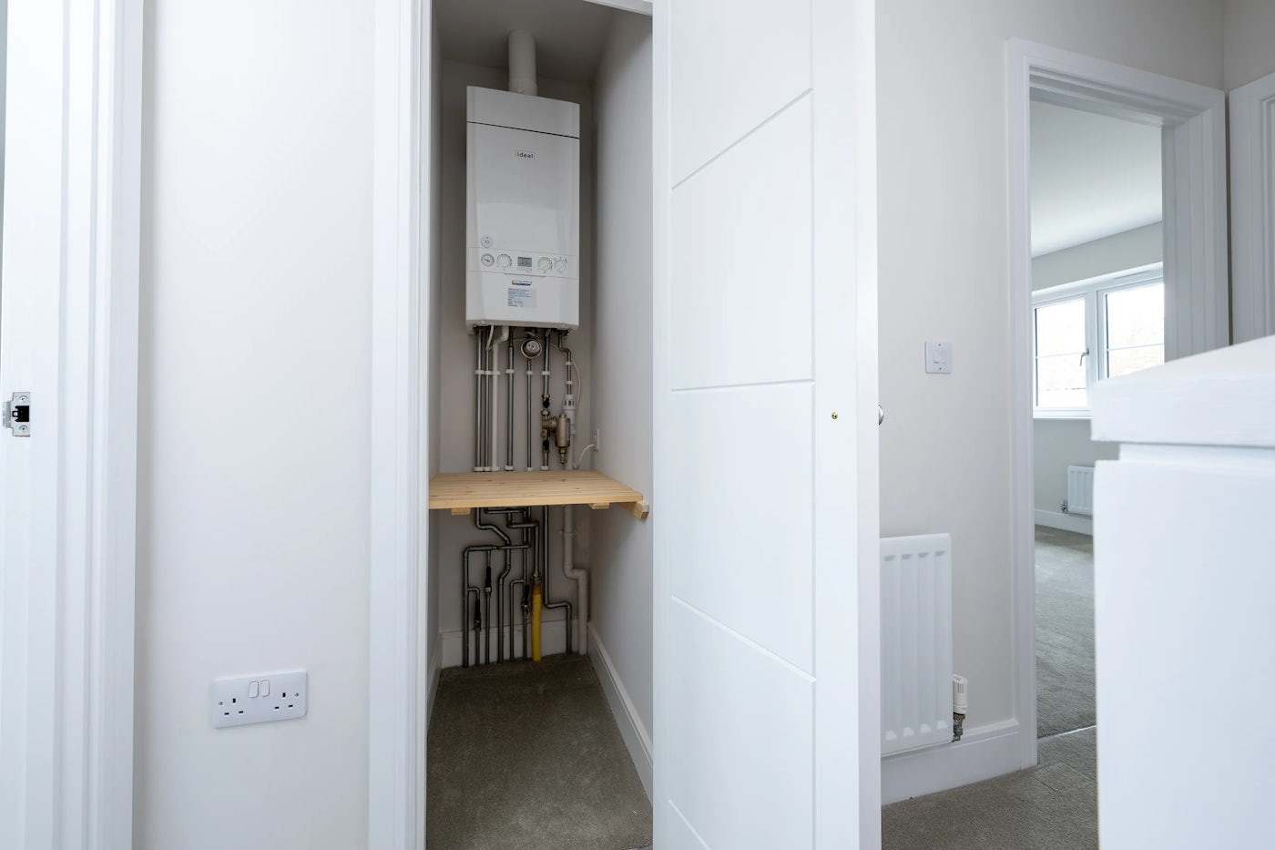 A utility closet with a wall-mounted boiler connected to exposed piping. Below the boiler is a wooden shelf, and the door is ajar, revealing a carpeted hallway with a radiator on the wall.