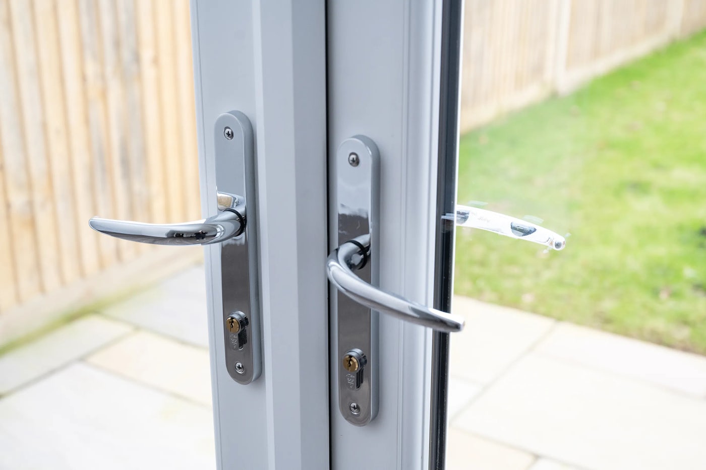 Close-up of modern metallic handles and locks on double glass doors, with a view of a garden and wooden fence visible through the glass.