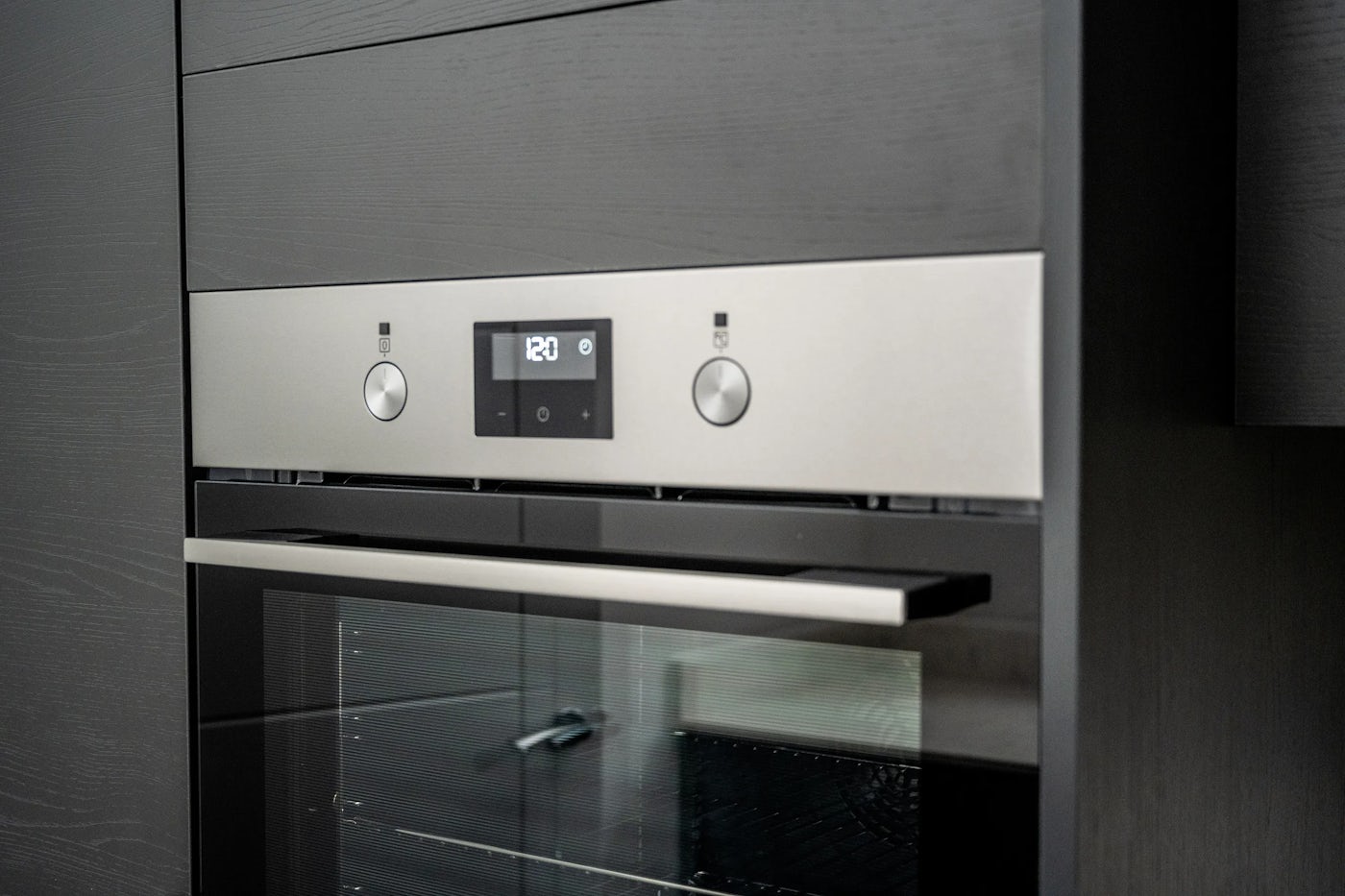 A built-in oven with a digital clock reading “12:01,” set within a dark-toned kitchen cabinet. The oven features a glass-front door, two control knobs with icons above them, and a minimalist design emphasizing clean lines and modern styling.