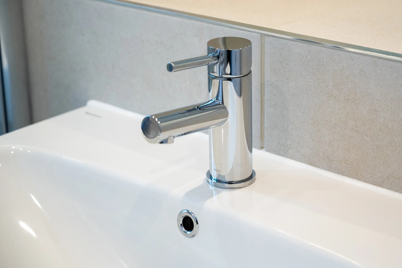 Close-up of a modern bathroom sink featuring a cylindrical chrome faucet with a single lever handle, set into a white basin with a chrome drain ring; a tiled wall and neutral countertop form the background.