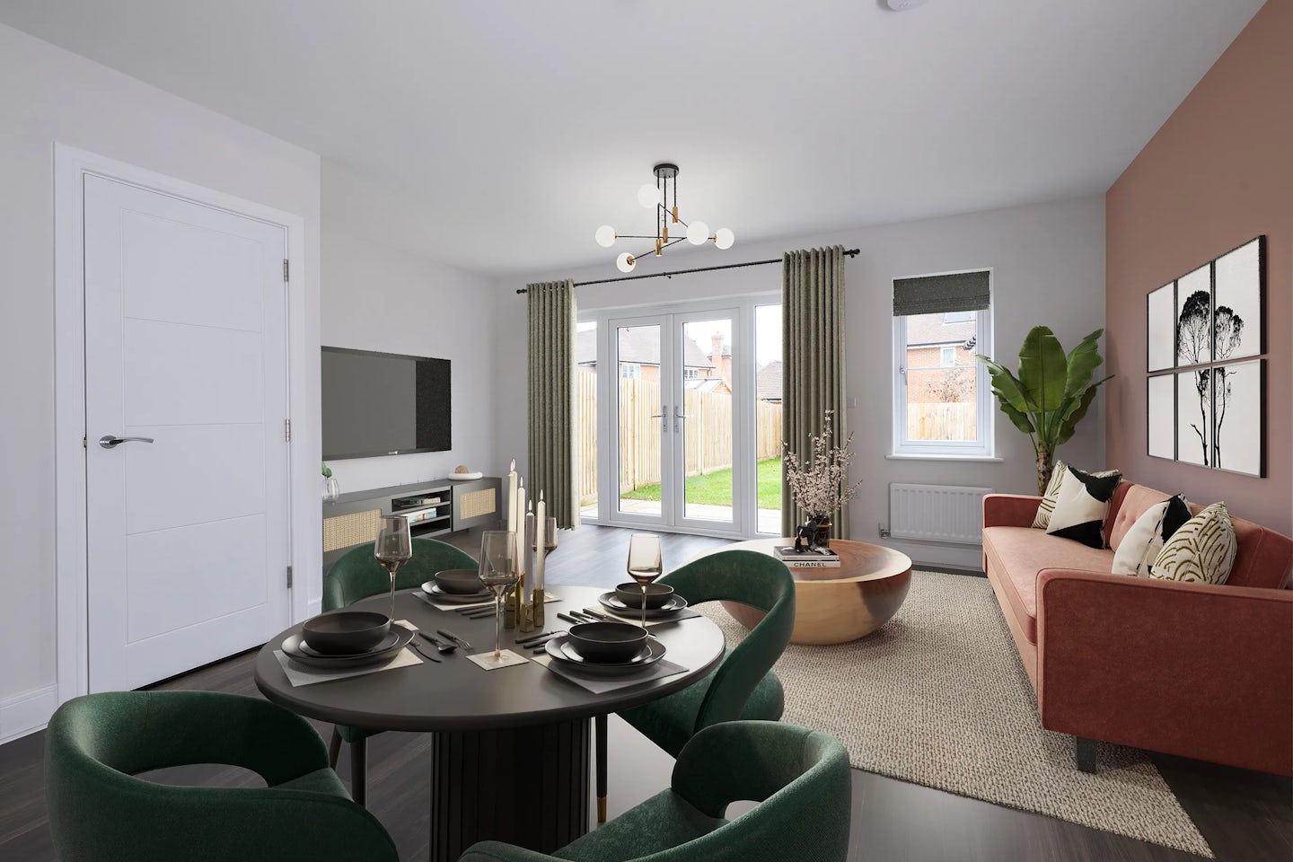Modern open-plan living and dining area with a red sofa, green dining chairs, and large glass doors overlooking a garden. A round black dining table is set with plates and candles, while a wooden coffee table and large plant add warmth. The room features white walls with a reddish-brown accent wall and is lit by a contemporary chandelier.