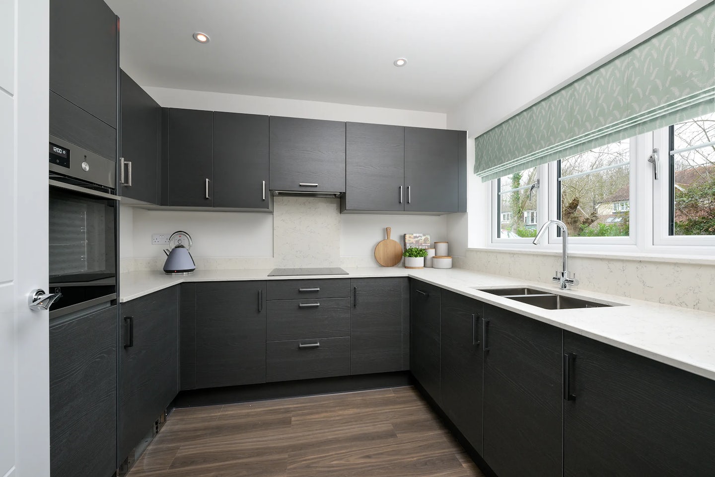A modern kitchen with dark grey cabinets, white countertops, and wooden flooring. A built-in oven is installed on the left, while a kettle and assorted kitchen items—like a cutting board, bowl, and containers—rest on the right-side counter. A large window with a green-patterned blind brings in natural light and offers an outdoor view, complementing the sleek, contemporary design.