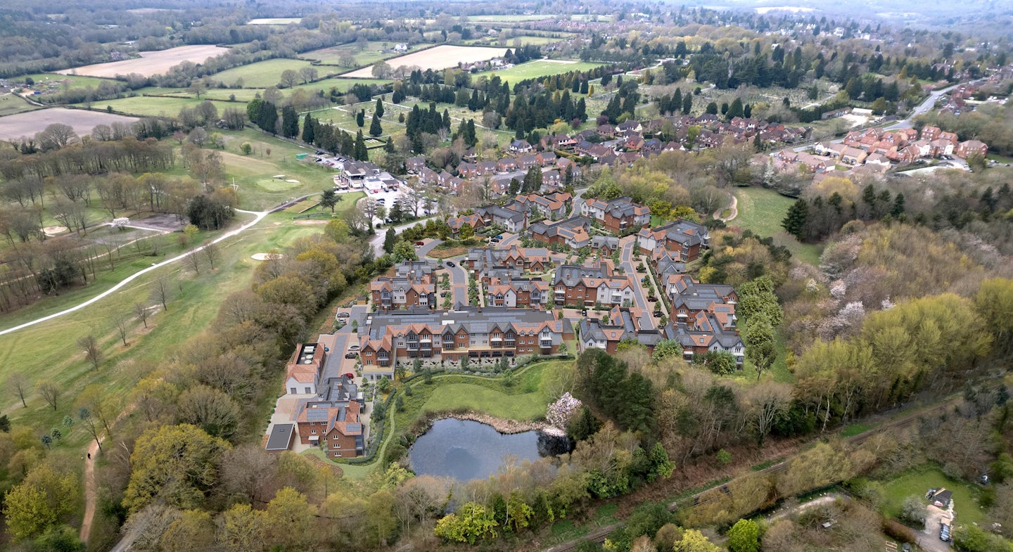 Aerial view of a planned residential area nestled within green surroundings. Rows of homes with brick facades and pitched roofs form an orderly neighborhood layout, bordered on the left by a sprawling golf course with manicured fairways and greens. A small pond, encircled by trees and vegetation, anchors the bottom centre of the image. The top of the frame reveals additional housing, open fields, and woodland, reflecting a seamless blend of built environment and nature.