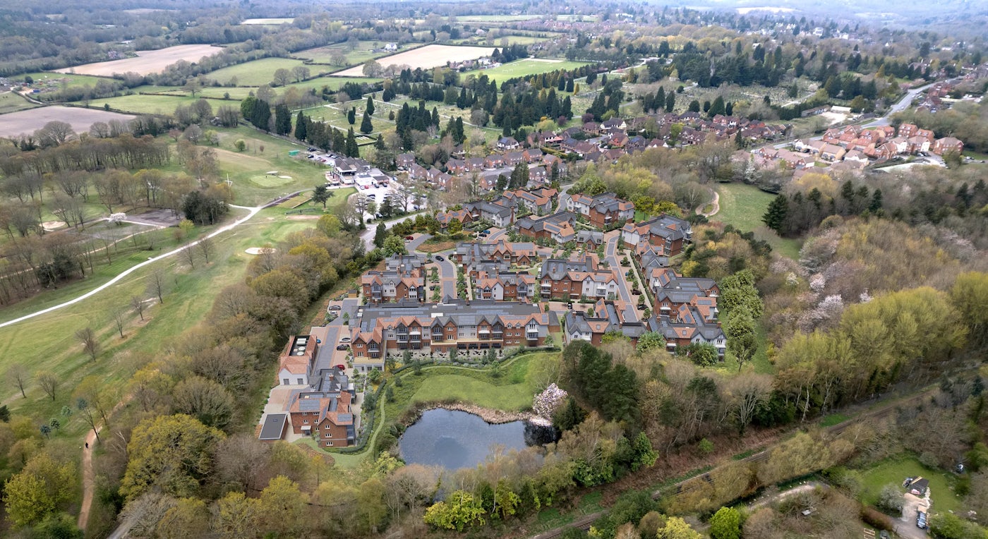 Aerial view of a planned residential area nestled within green surroundings. Rows of homes with brick facades and pitched roofs form an orderly neighborhood layout, bordered on the left by a sprawling golf course with manicured fairways and greens. A small pond, encircled by trees and vegetation, anchors the bottom centre of the image. The top of the frame reveals additional housing, open fields, and woodland, reflecting a seamless blend of built environment and nature.