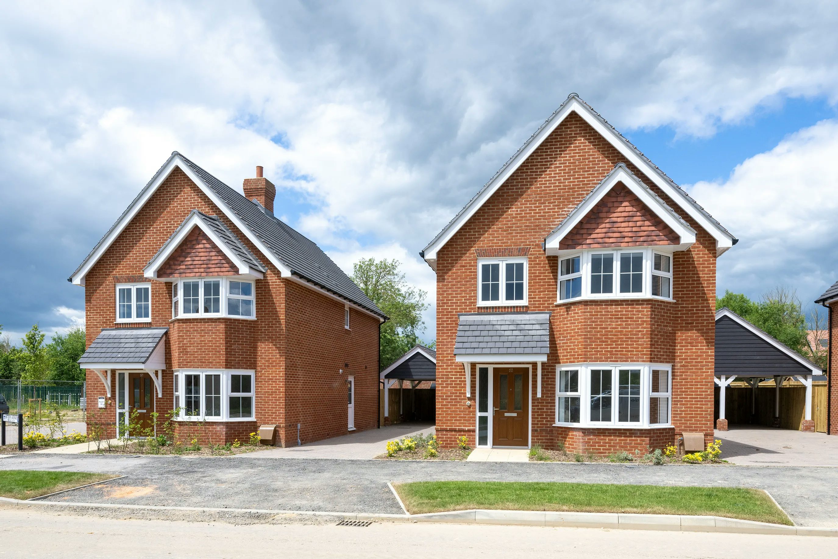 Two contemporary detached houses with red brick exteriors and pitched, dark grey roofs, positioned side by side along a paved driveway. Each has white-framed windows and doors; the left house features a chimney. Neatly maintained greenery borders the path, and a partly cloudy sky stretches overhead.