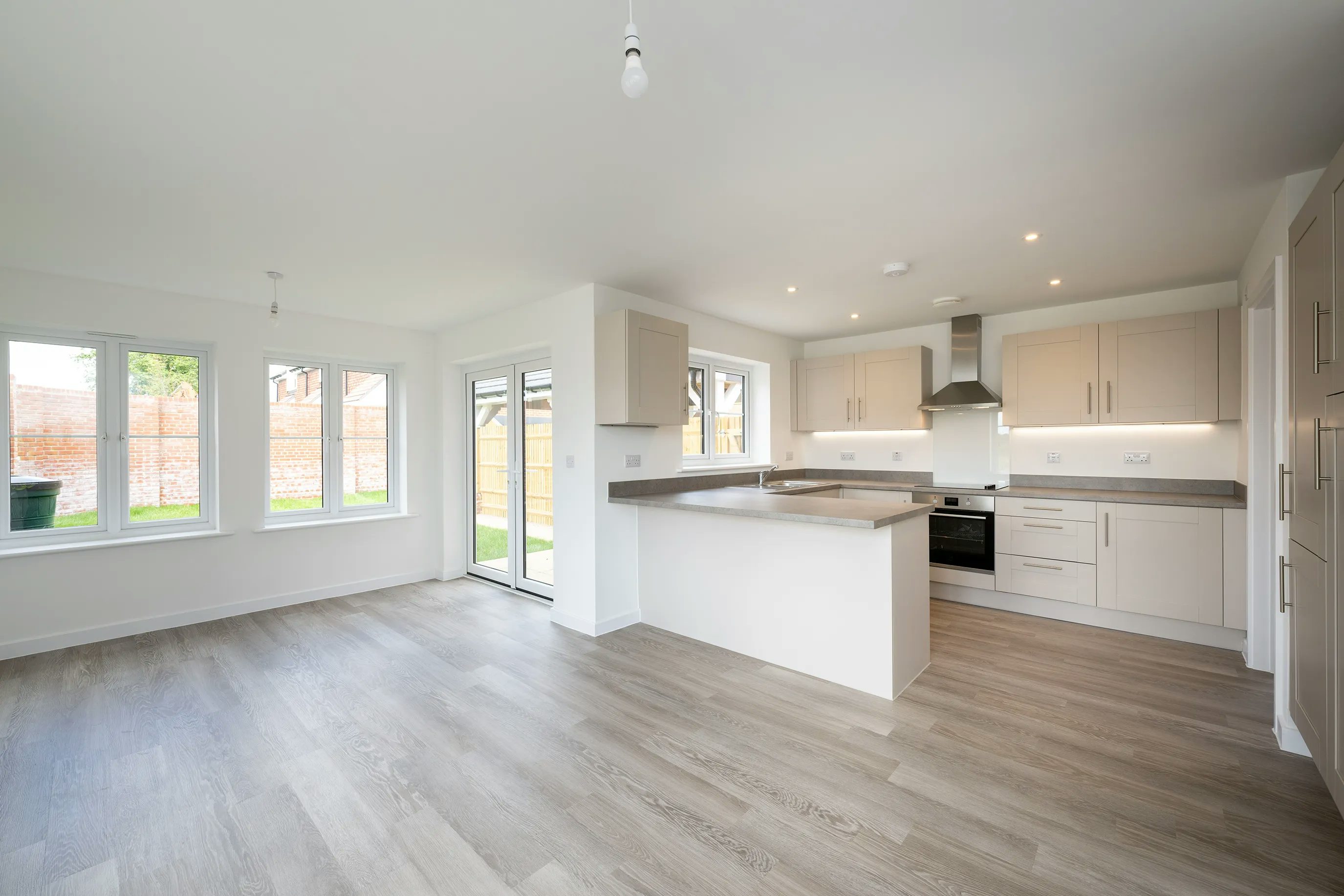 Modern open-plan kitchen and living area with white cabinetry, stainless steel appliances, and a central island topped with a light countertop; large windows flood the space with natural light, highlighting the light wood laminate flooring.