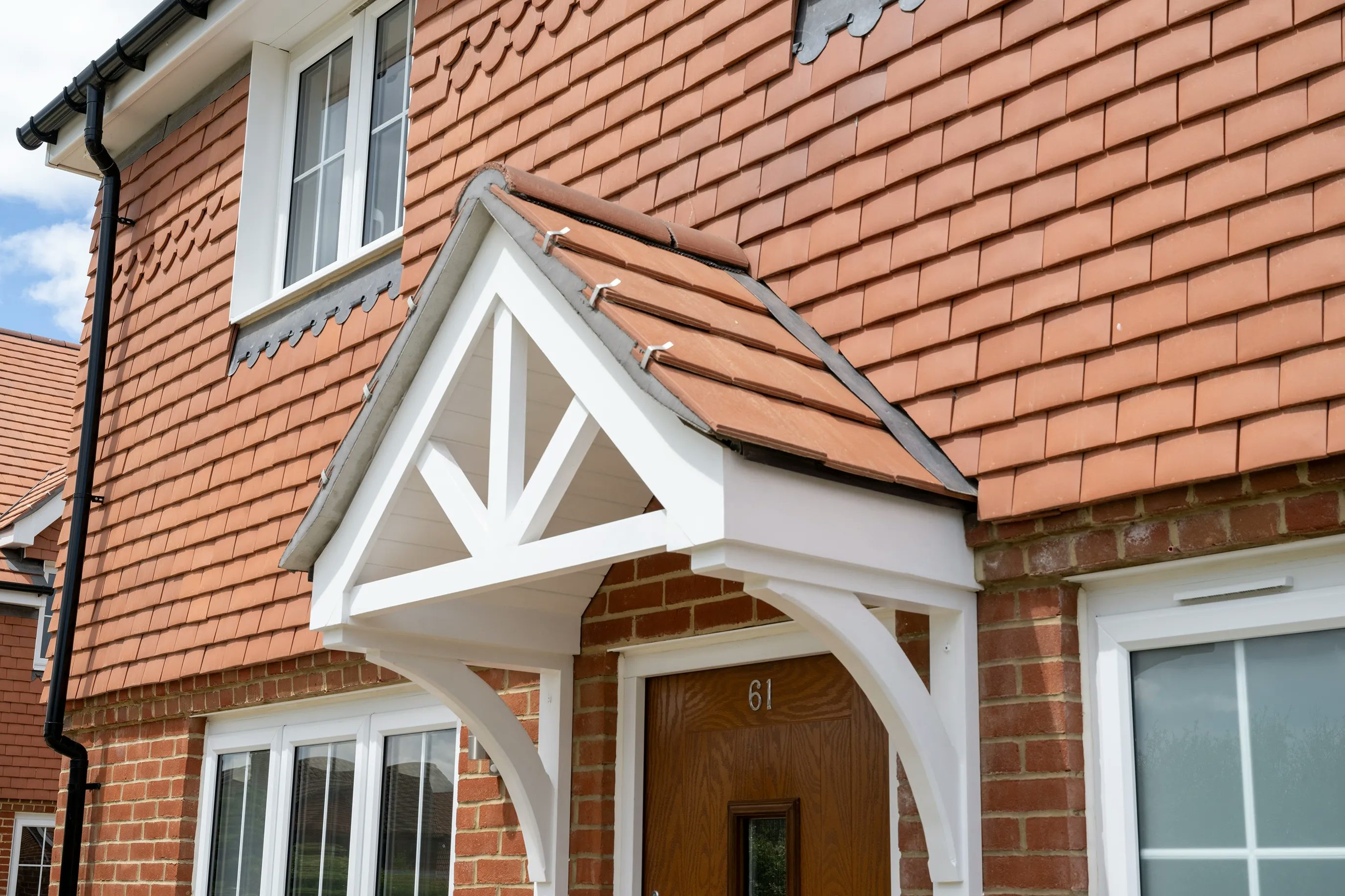 Front entrance of a red brick house with a white wooden porch and red tiled roof. The door, numbered 61, is centered beneath a small gabled porch with decorative trim. White-framed windows flank the entrance, complementing the clean, traditional aesthetic.