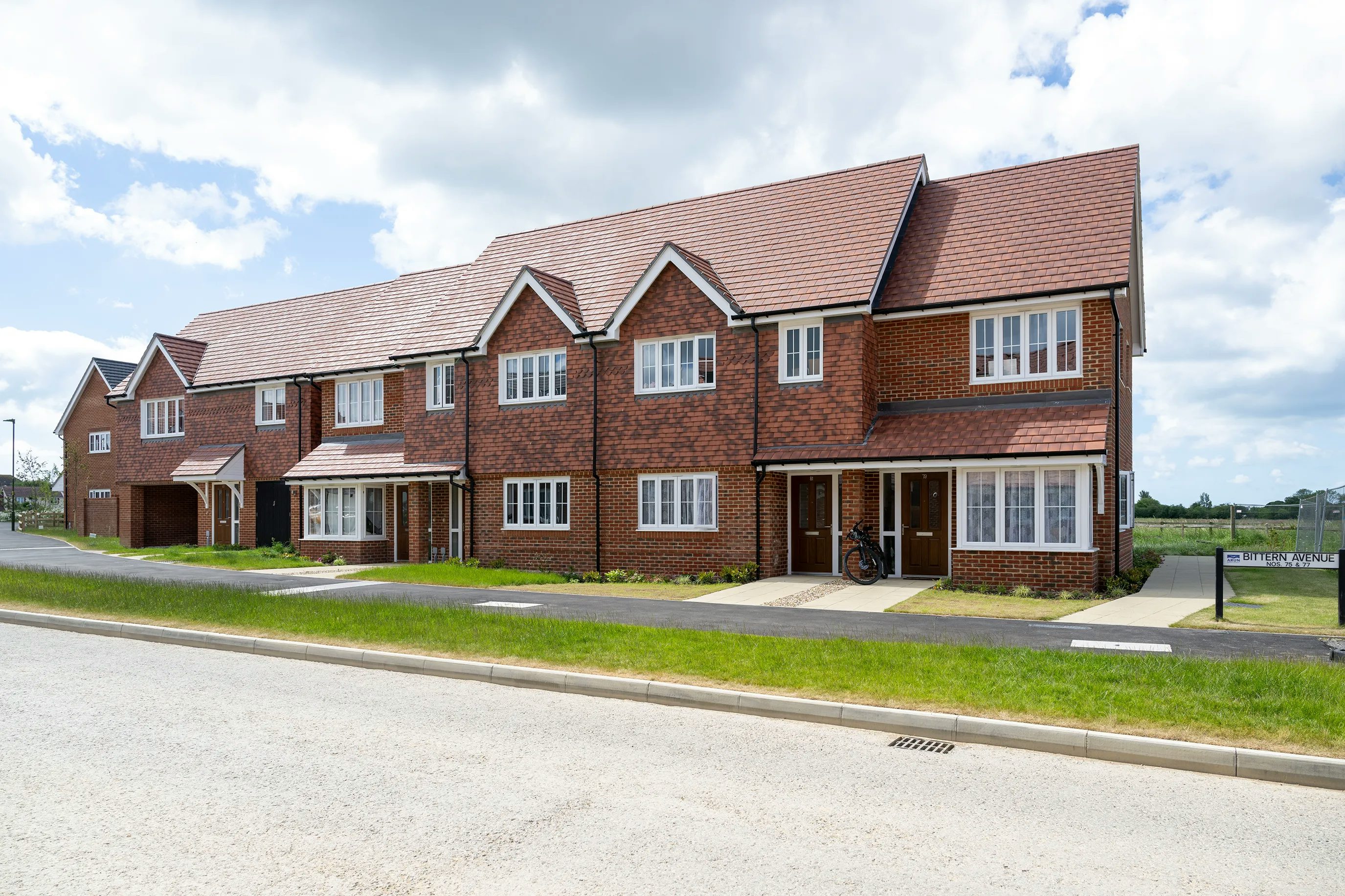 Row of newly built red-brick townhouses with white-framed windows and pitched roofs, lining a paved road with a grassy verge and a bicycle parked out front under a partly cloudy sky.