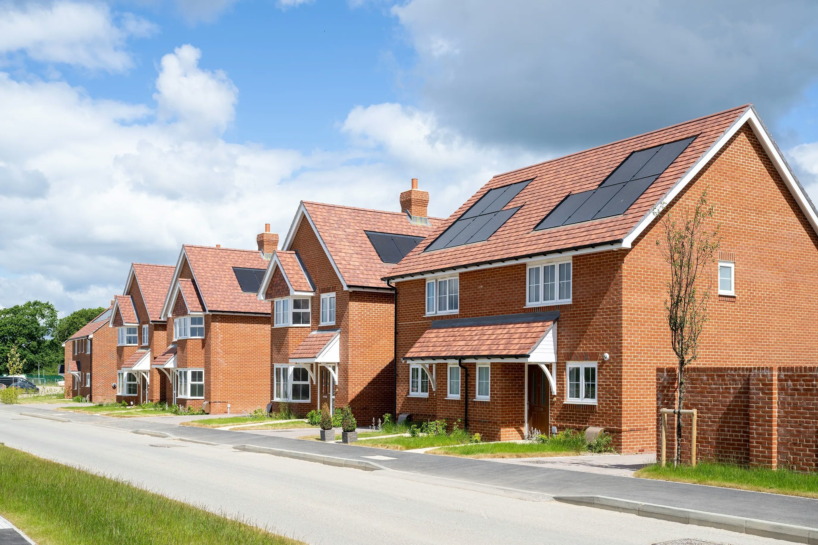 A row of contemporary brick houses lines a quiet residential street, each featuring a pitched roof fitted with solar panels. The homes are set back behind small green lawns, and a paved road runs parallel to the row. Overhead, the sky is partly cloudy, casting soft light across the scene.