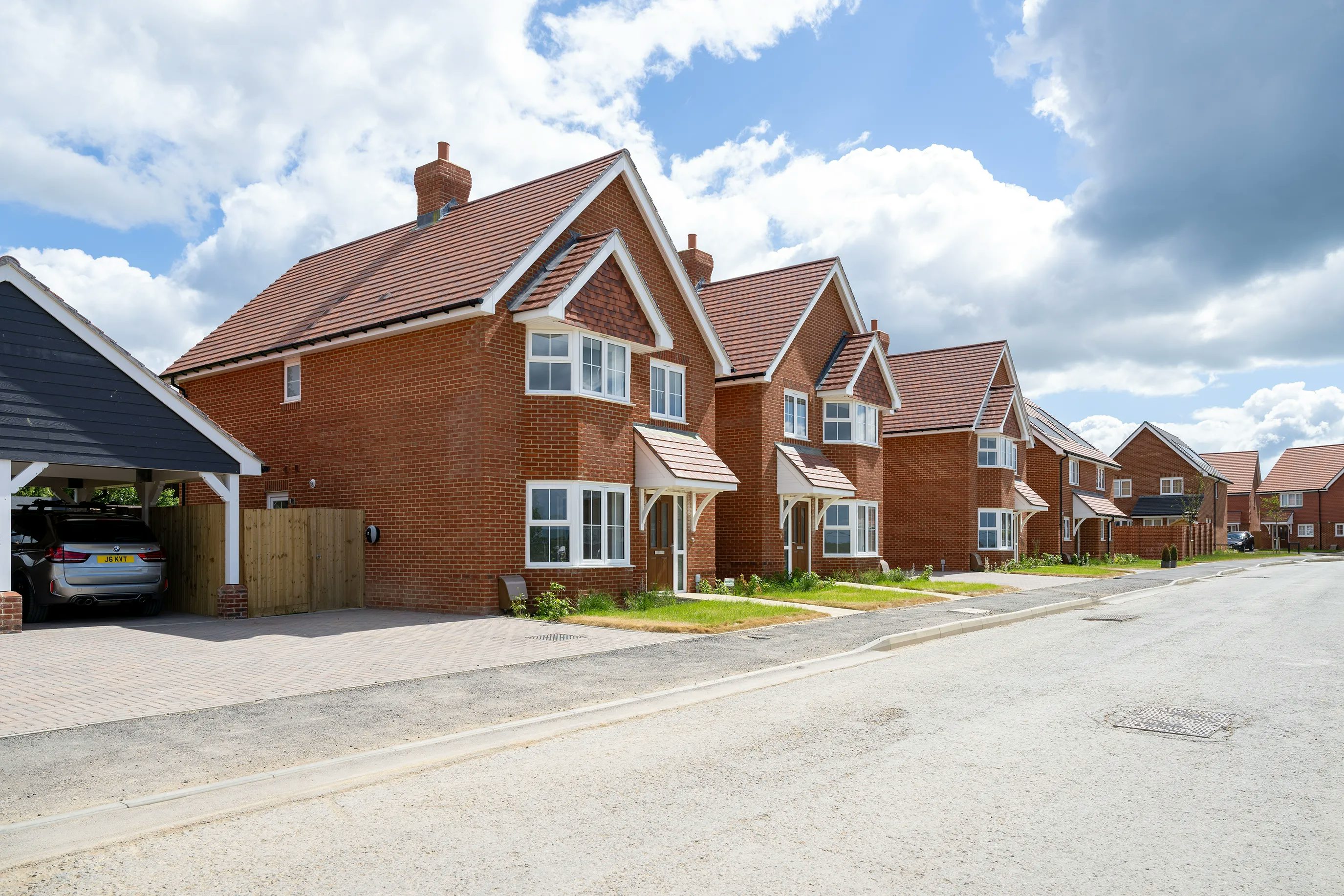 A row of modern red-brick detached houses with pitched roofs lines a quiet suburban street. Each home features a small front garden and a private driveway; one driveway holds a parked car. The sky is bright with scattered clouds, suggesting a clear, pleasant day. The overall atmosphere is calm and residential.