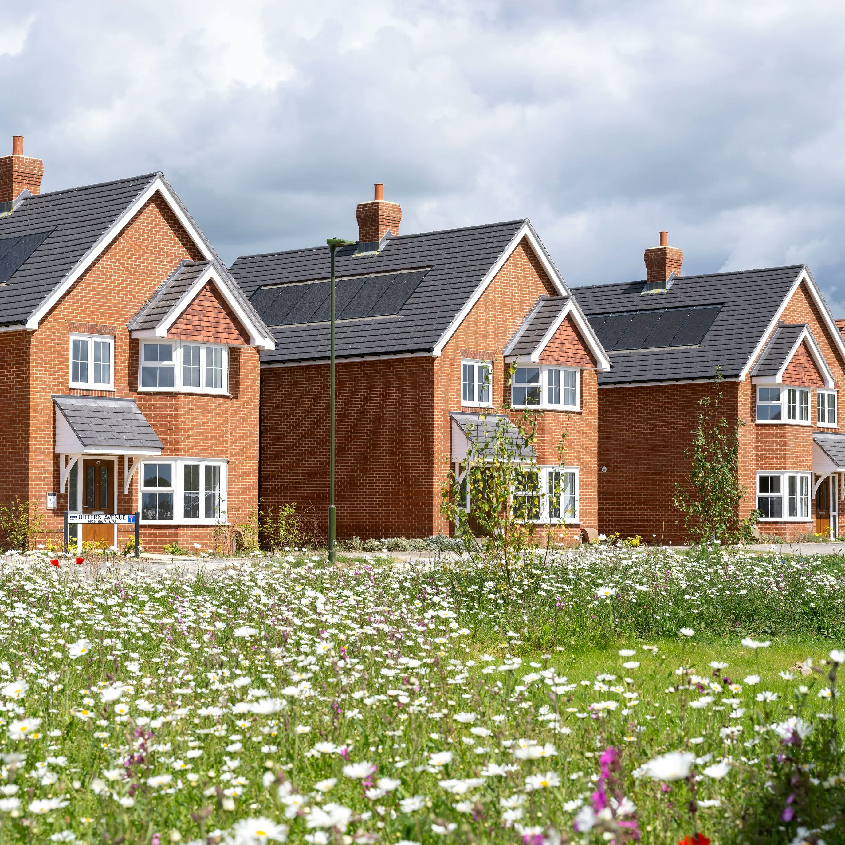 Three contemporary brick houses with steeply pitched roofs and front porches sit beyond a colorful wildflower field under a partly cloudy sky, creating a serene and idyllic residential scene.