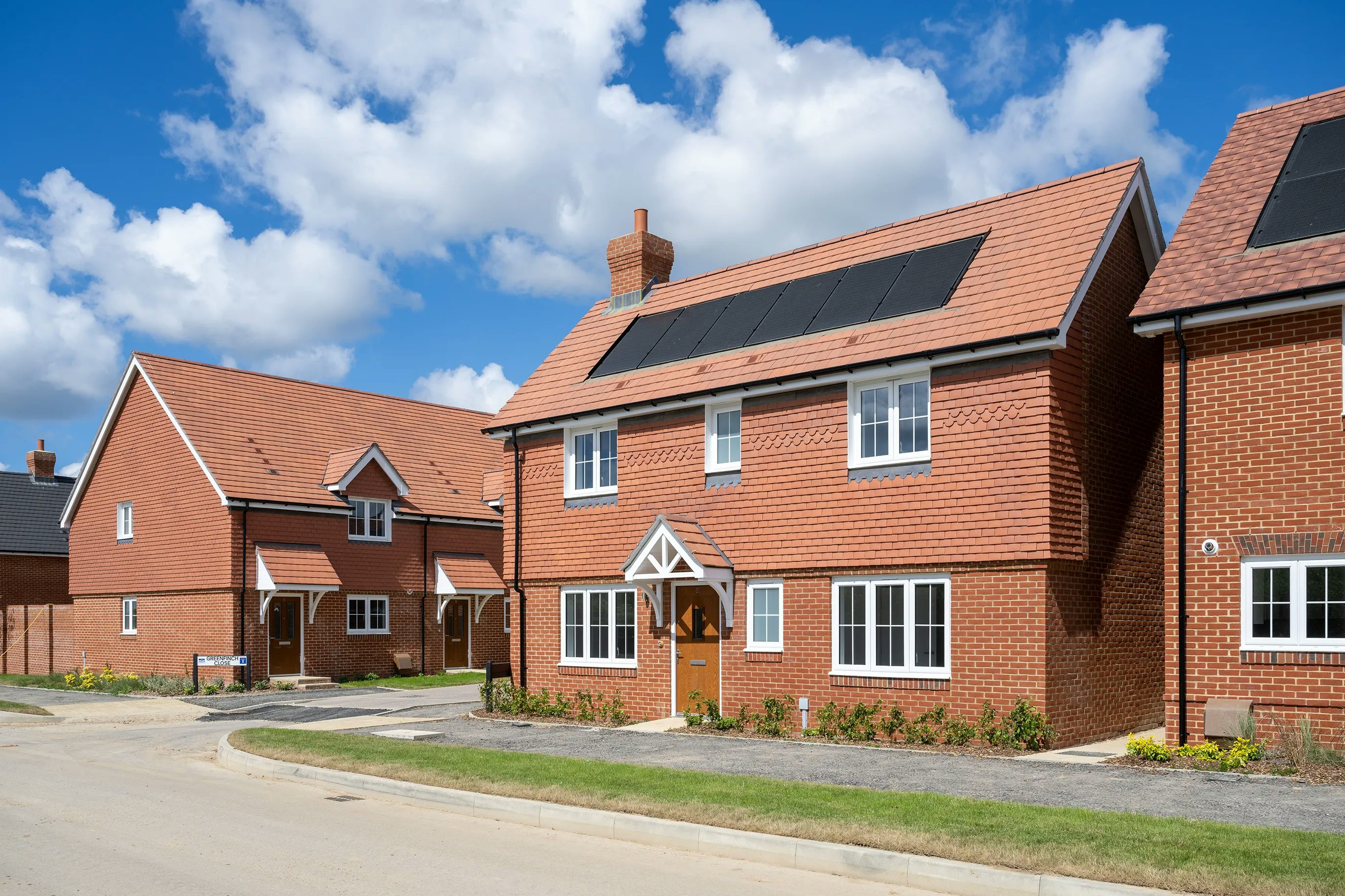 A row of contemporary detached homes with red-tiled roofs and brick facades, each featuring solar panels integrated into the roof design. The houses are evenly spaced along a clean, quiet street under a bright blue sky with scattered white clouds, reflecting a focus on sustainability within a modern suburban setting.