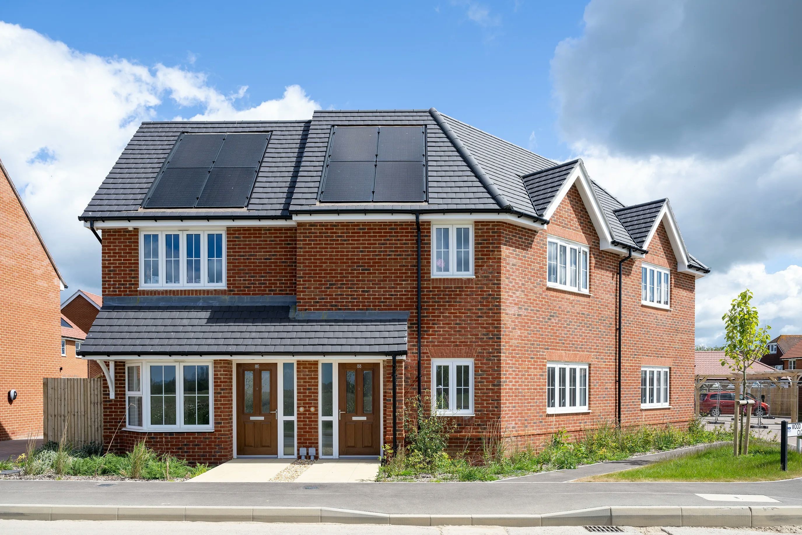 Semi-detached brick house in a suburban neighborhood with a tiled roof and solar panels. Two white-framed doors suggest separate units, complemented by white-framed windows. A small front garden features green plants and a leafy tree under a partly cloudy sky.