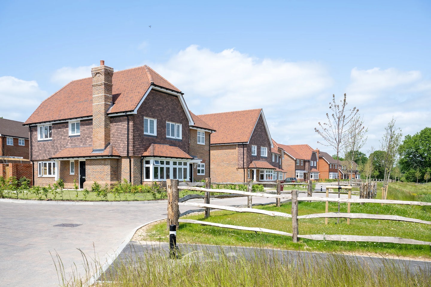 A row of modern suburban houses with red-tiled roofs and brick facades lines a gently curved residential street. A wooden fence runs parallel to the paved road, separating the homes from a well-kept grassy verge. Trees and greenery are scattered throughout the scene beneath a mostly clear sky with a few light clouds.