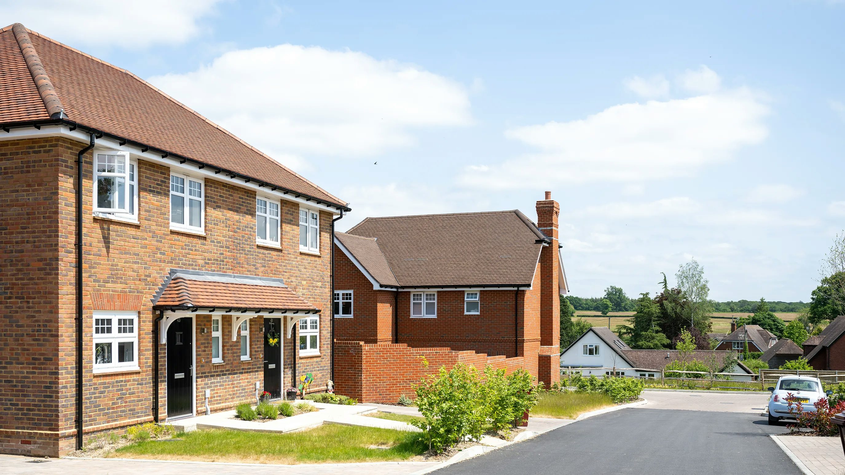 A two-story brick house with white-framed windows and a black front door, set amid a tidy front garden with trimmed grass and shrubs. To the left is a neighboring house with a chimney, and in the background, more suburban homes are framed by green trees and a bright blue sky with scattered clouds. A parked car sits along the curb on the right, reinforcing the residential, peaceful atmosphere.