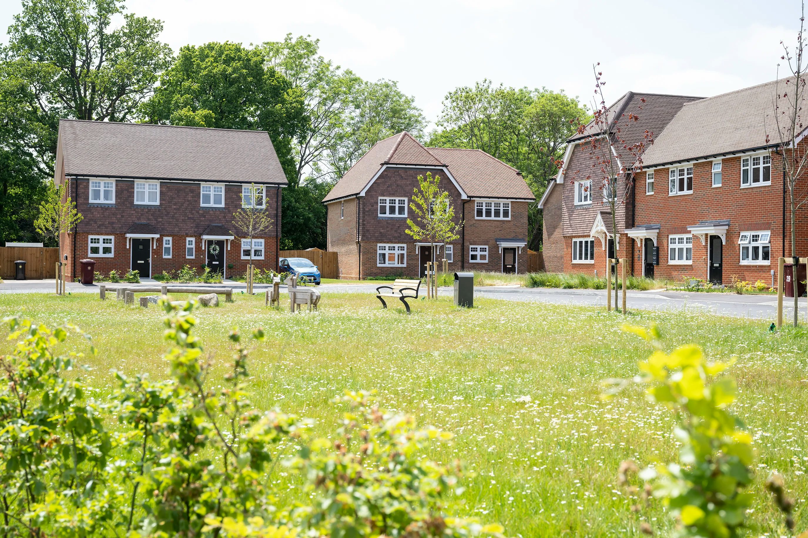 A quiet suburban street lined with two-story red-brick houses, each featuring white-framed windows and gable roofs. In the foreground, a small communal green space includes neatly mown grass, a few leafy trees, and wooden benches. Several cars are parked along the paved road, adding to the everyday residential atmosphere. The overall mood is peaceful and well-kept, with a focus on community and liveability.