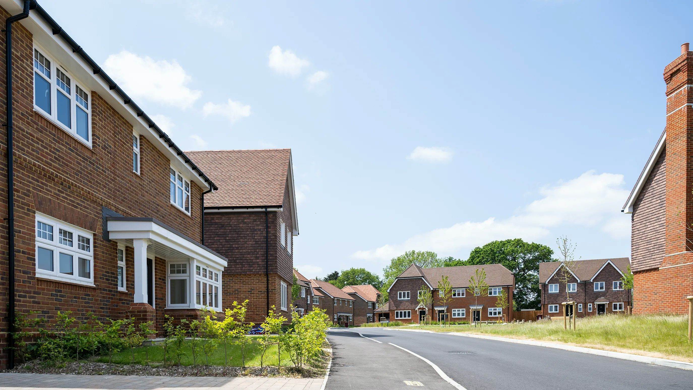 Curved residential street lined with red-brick houses featuring white-framed windows. A paved road and sidewalk run alongside low greenery and small plants. The sky is mostly clear with a few scattered clouds, suggesting a sunny day.