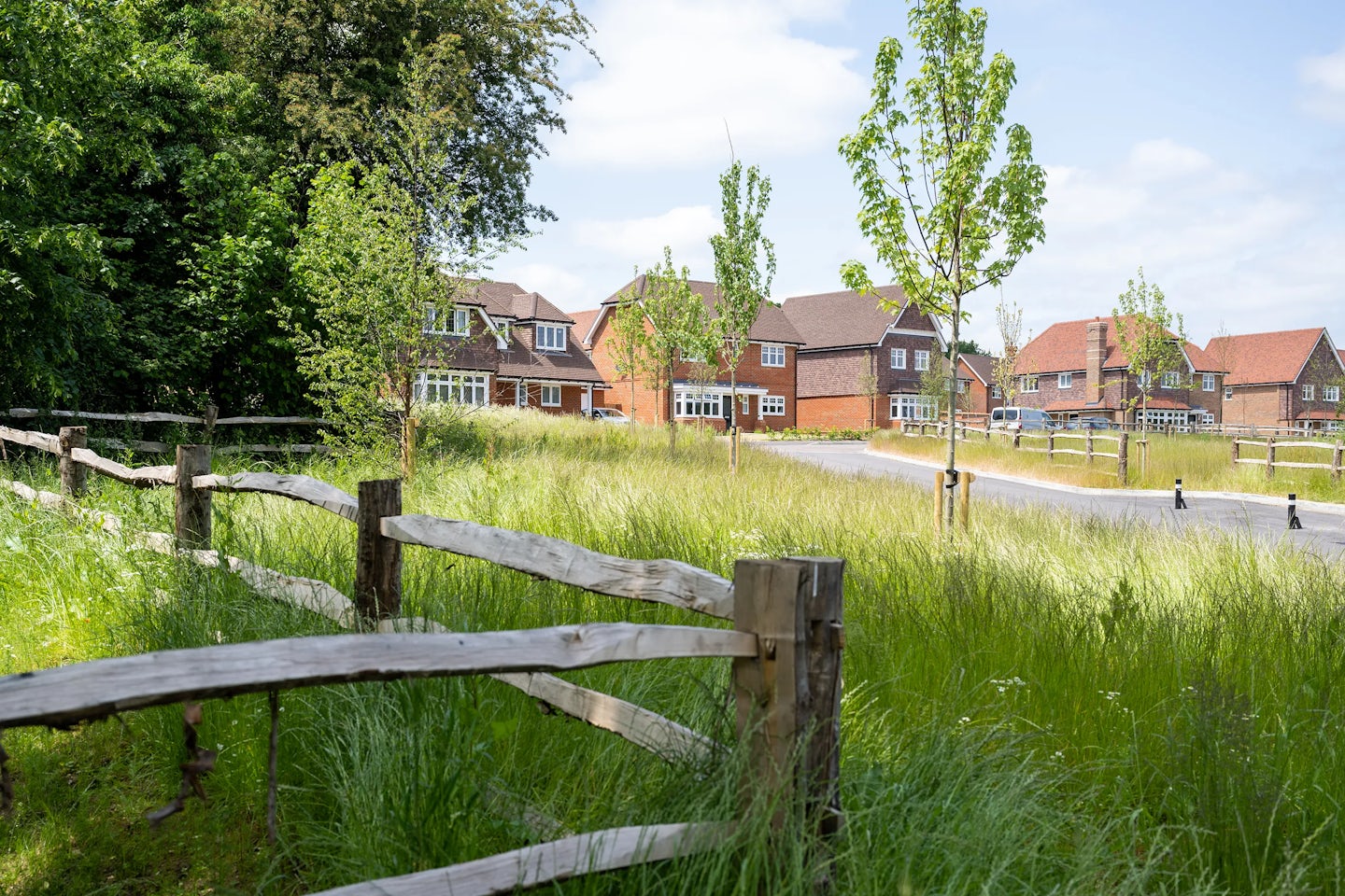 A scenic suburban street curves gently past a row of brick houses with tiled roofs, bordered by a low wooden fence in the foreground. Tall grass and mature trees line the roadside, contributing to a peaceful, green atmosphere. The neighborhood appears quiet and well-kept, bathed in soft daylight that enhances its calm, residential charm.