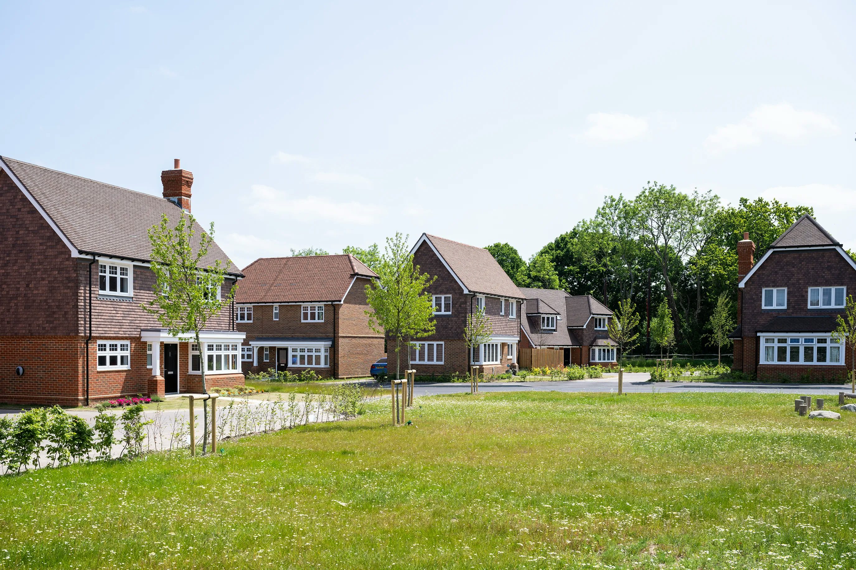 A row of neatly kept brick houses in a suburban neighborhood, each with pitched roofs and chimneys, set against a clear blue sky. The scene features manicured lawns, young trees lining the street, and no visible people or vehicles, creating a calm, residential atmosphere.