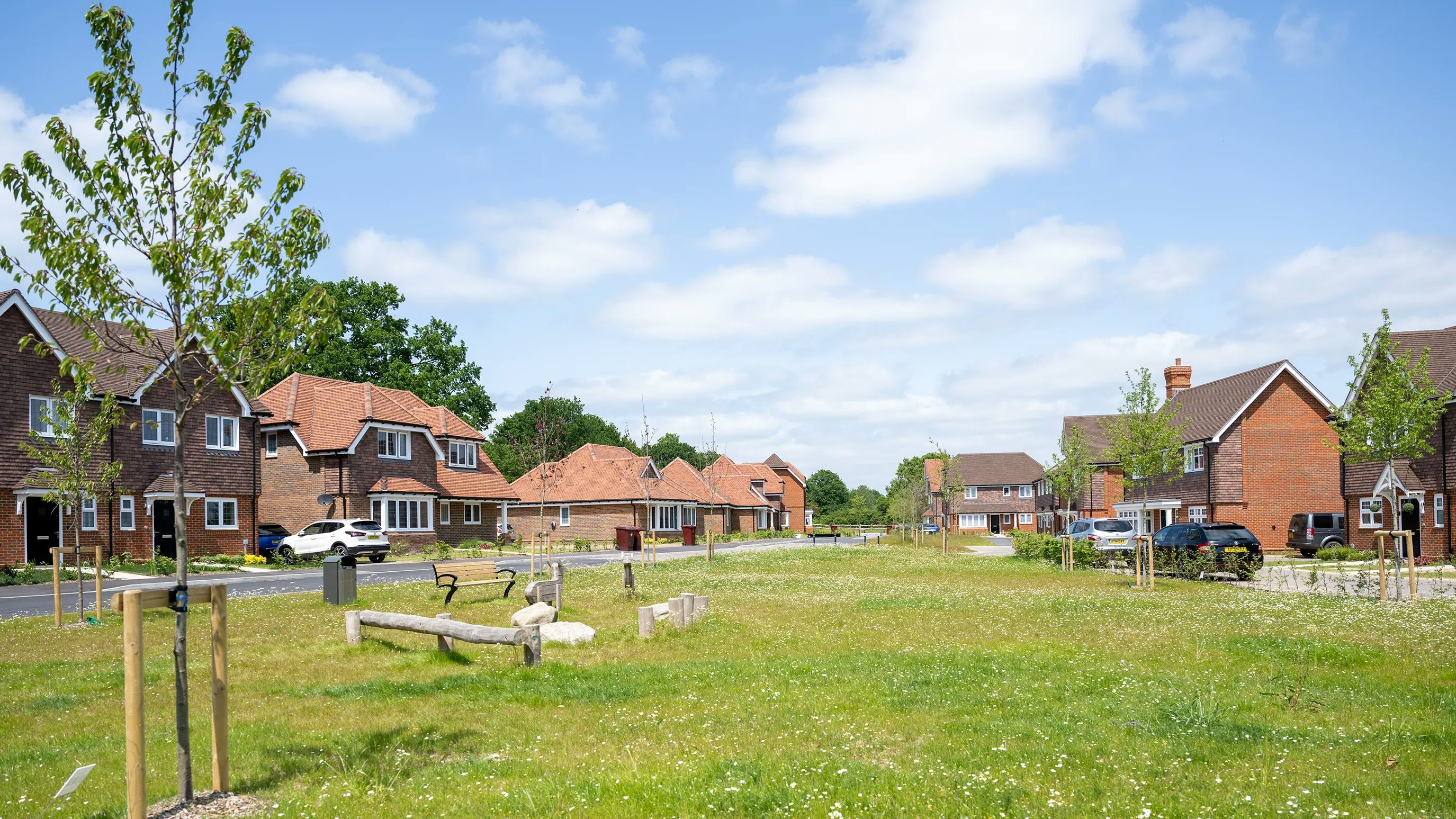 A peaceful suburban street scene with red-brick homes, neatly trimmed lawns, and young trees lining the sidewalk. A wooden playground set—featuring a slide and swings—sits on a grassy patch, suggesting a family-friendly environment. Several parked cars dot the roadside under a blue sky with light cloud cover, lending a calm and inviting atmosphere.