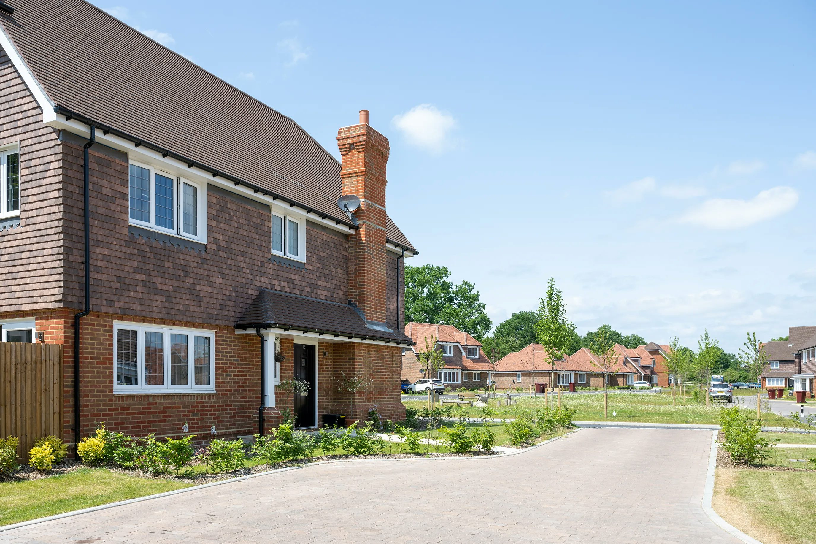 A two-story brick house with a dark brown roof and white-trimmed windows, set in a neatly landscaped suburban neighborhood. The front garden features low shrubs and plants, while a paved driveway curves past the house and leads to neighboring homes under a bright, mostly clear sky.