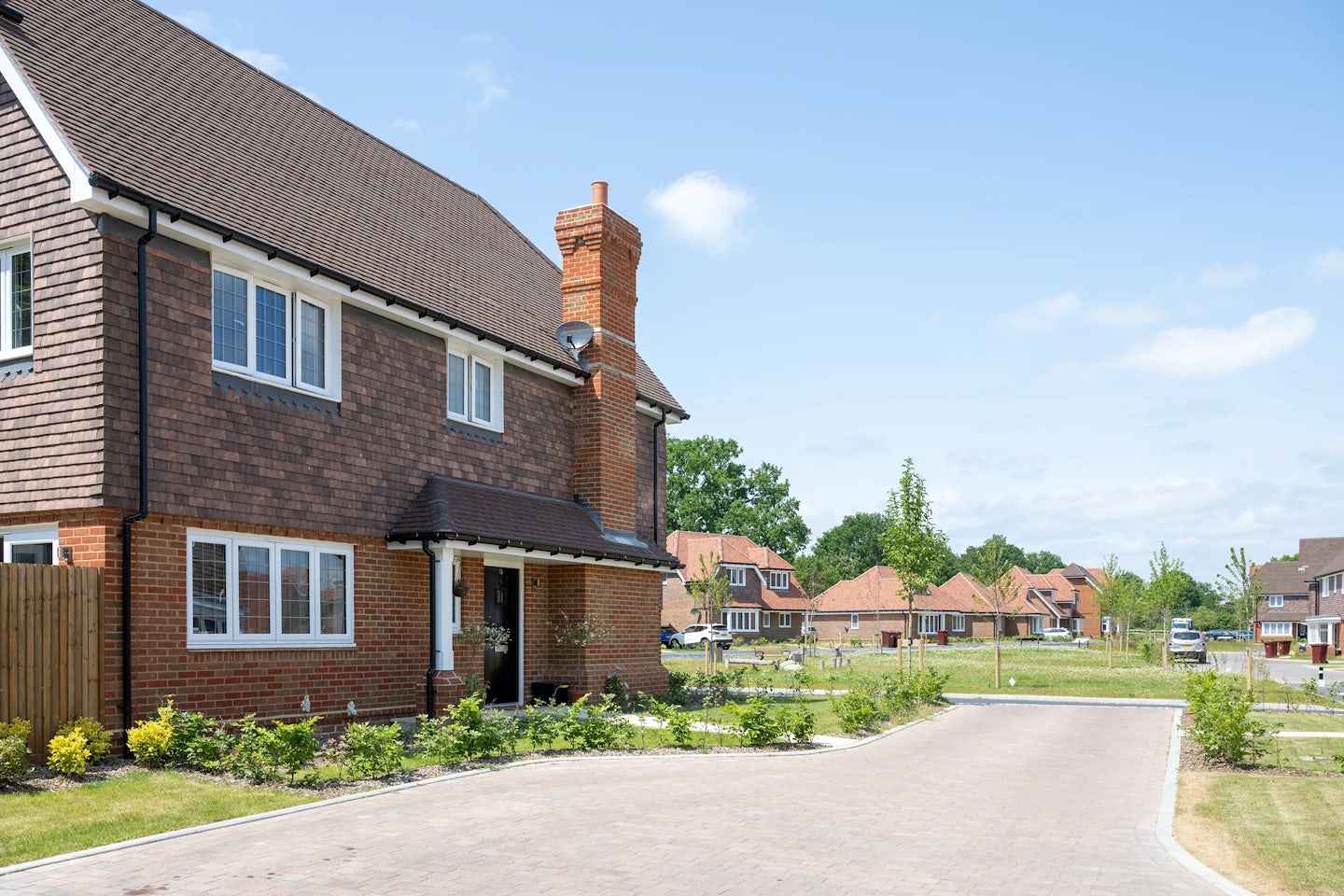 A two-story brick house with a dark brown roof and white-trimmed windows, set in a neatly landscaped suburban neighborhood. The front garden features low shrubs and plants, while a paved driveway curves past the house and leads to neighboring homes under a bright, mostly clear sky.