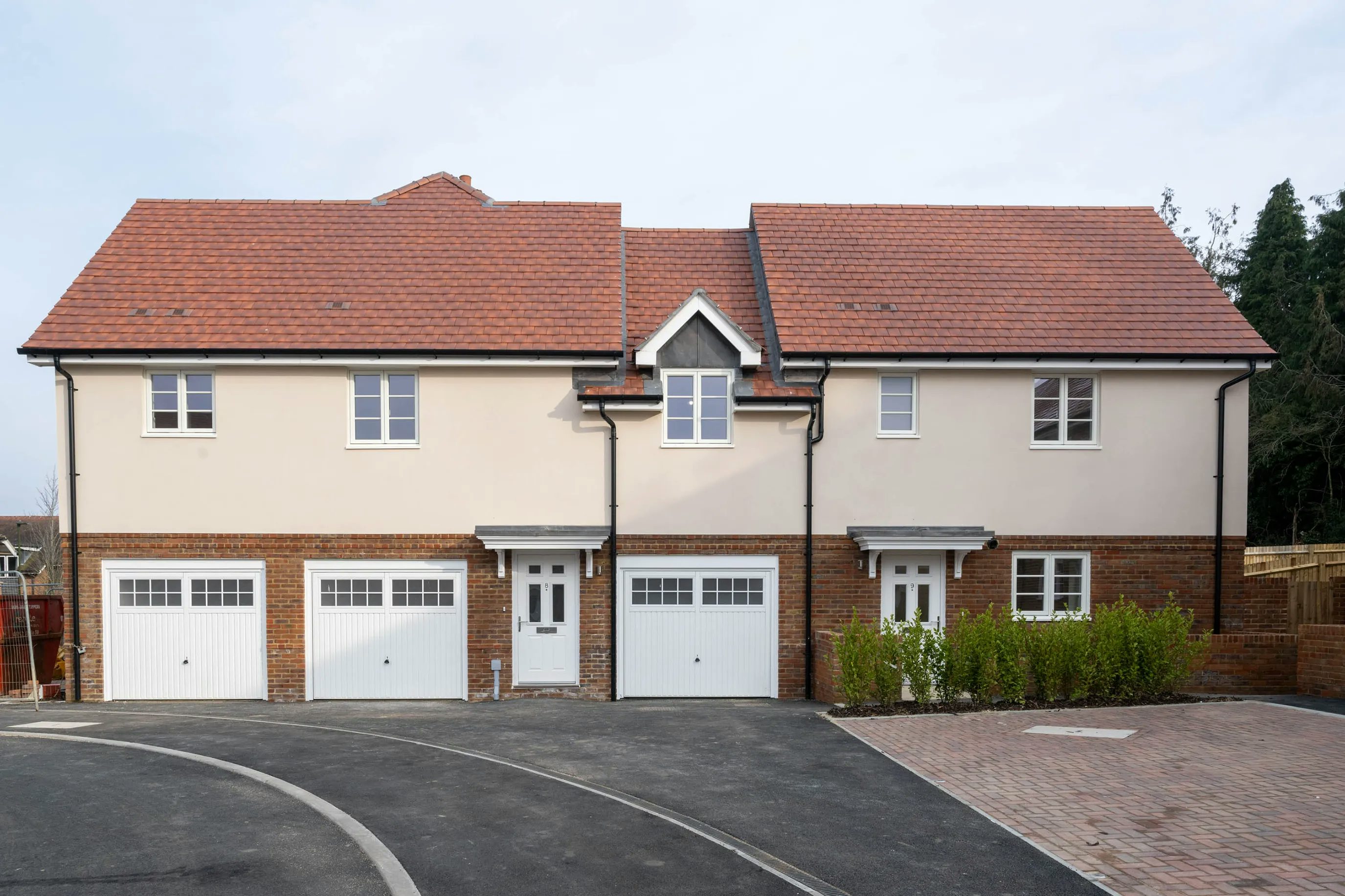 A row of two modern townhouses with red-tiled roofs and cream-colored exteriors, each featuring a garage with a white door, upper and lower front-facing windows, and a small gable accent on the middle unit. The driveways are paved, and the rightmost home includes a small brick-paved entry area with greenery.