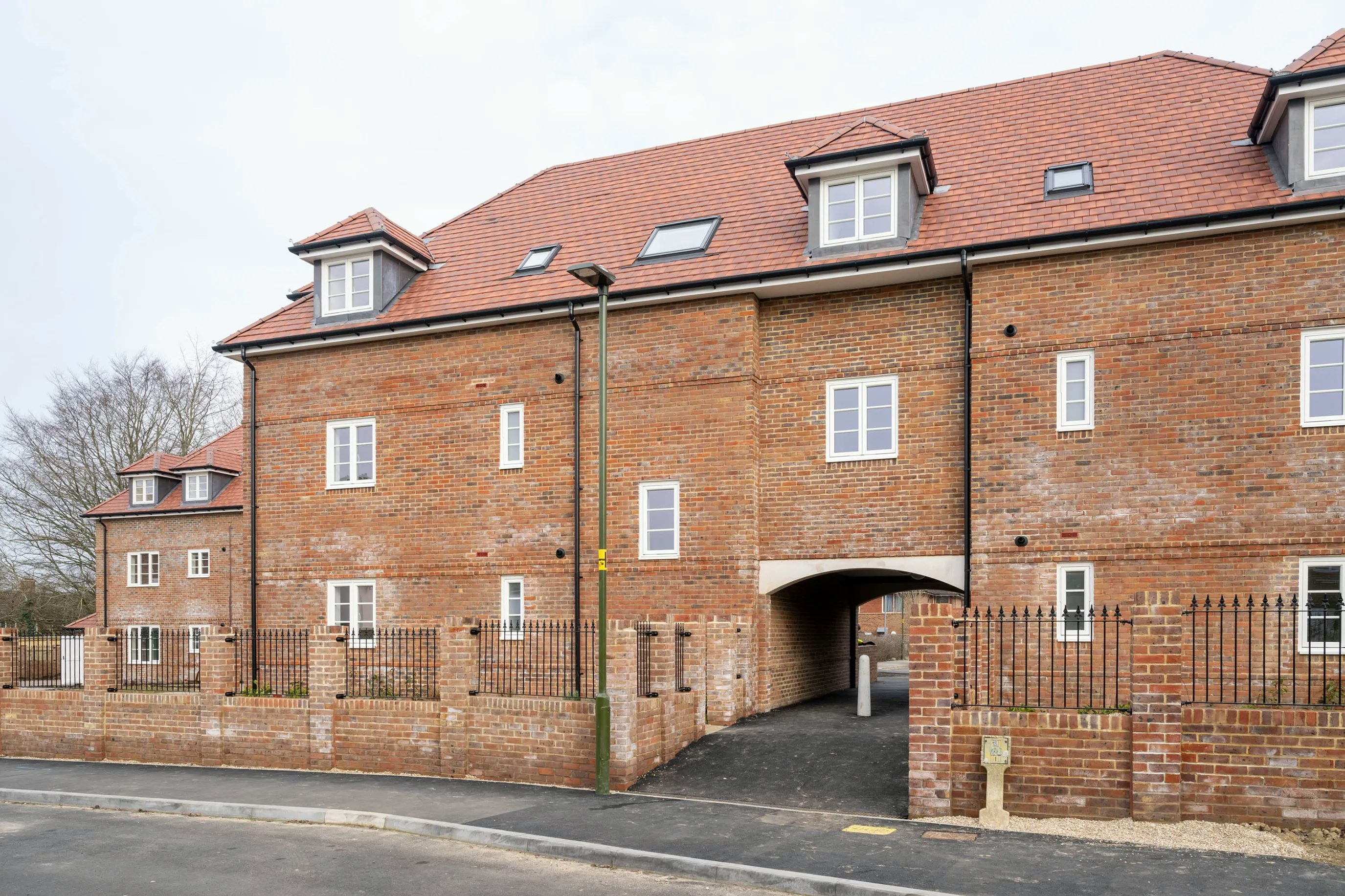 A modern, multi-story brick building with a red-tiled roof and several dormer windows. The structure features an archway at its center leading to a covered passage. It’s enclosed by a metal and brick fence, with a paved sidewalk and street in the foreground.