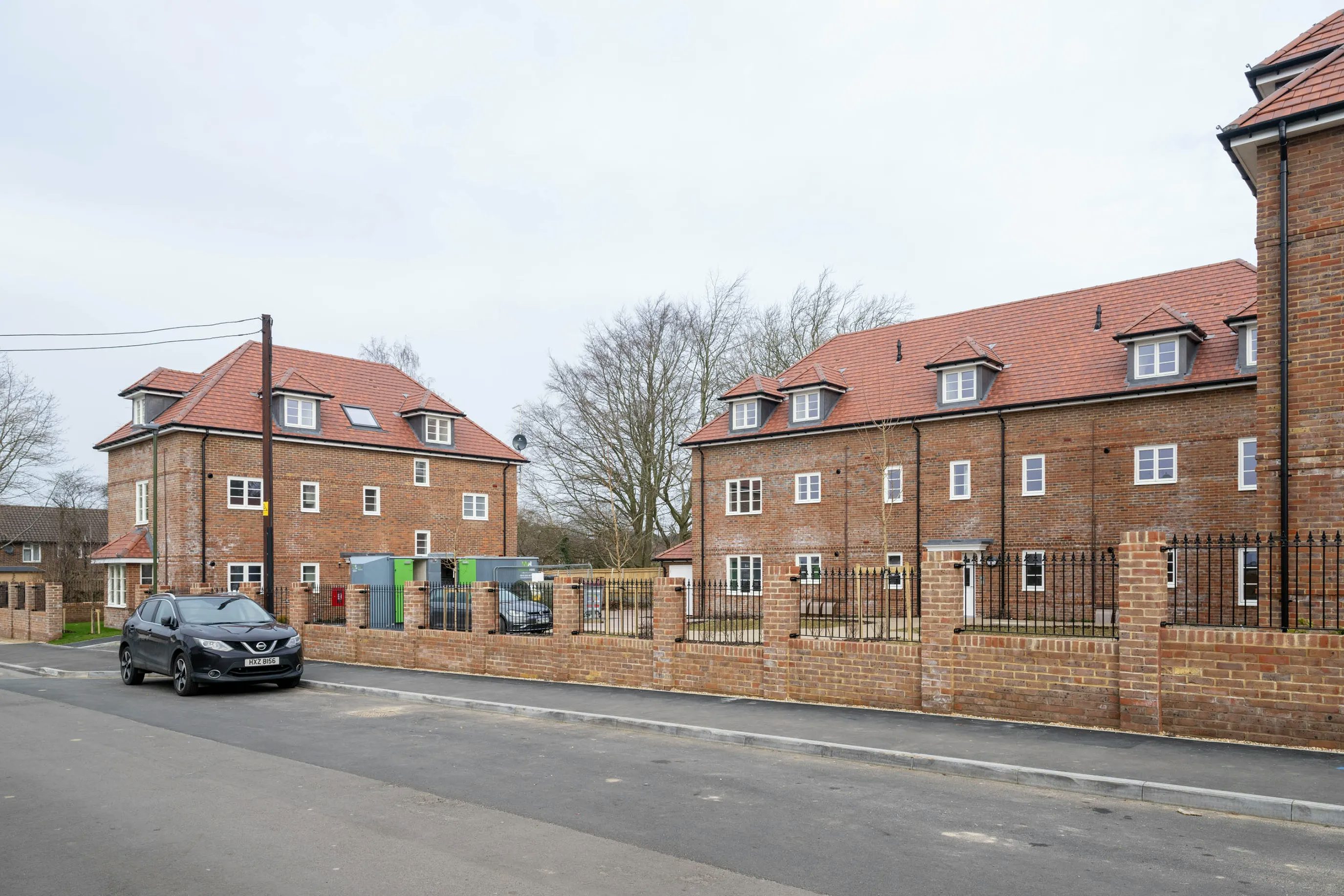 A quiet residential street featuring three newly built multi-story brick houses with red-tiled roofs and dormer windows. A black car is parked at the curb in front of the houses, which are enclosed by a low brick wall topped with black metal railings. Mature trees rise behind the homes, and the scene unfolds under an overcast sky, suggesting a calm, subdued atmosphere.