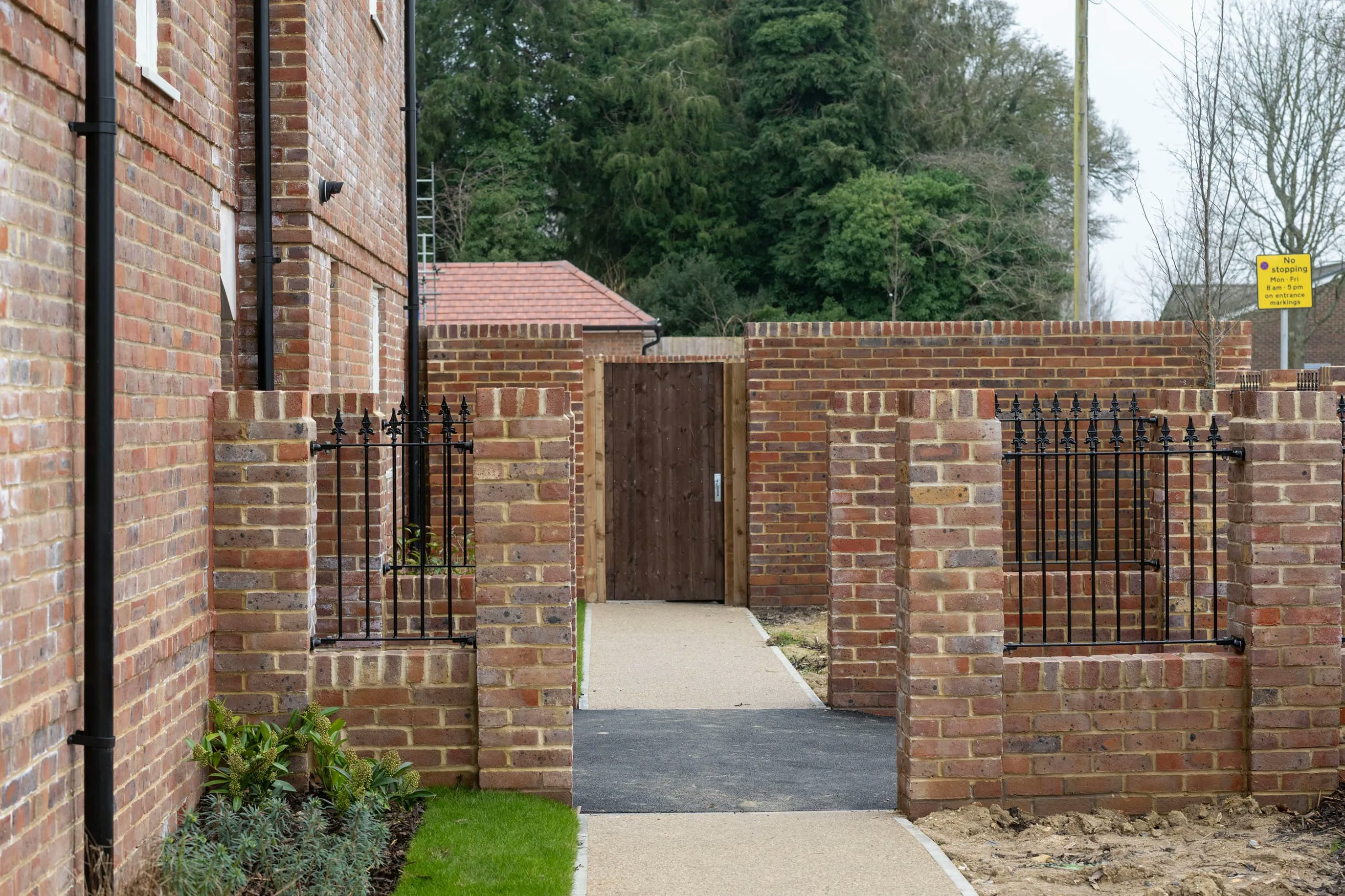 A brick pathway bordered by low brick walls and black railings leads to a wooden gate. Lush greenery peeks over the left side, while a yellow sign is visible on the right. In the distance, a red-roofed building is partially obscured by trees.