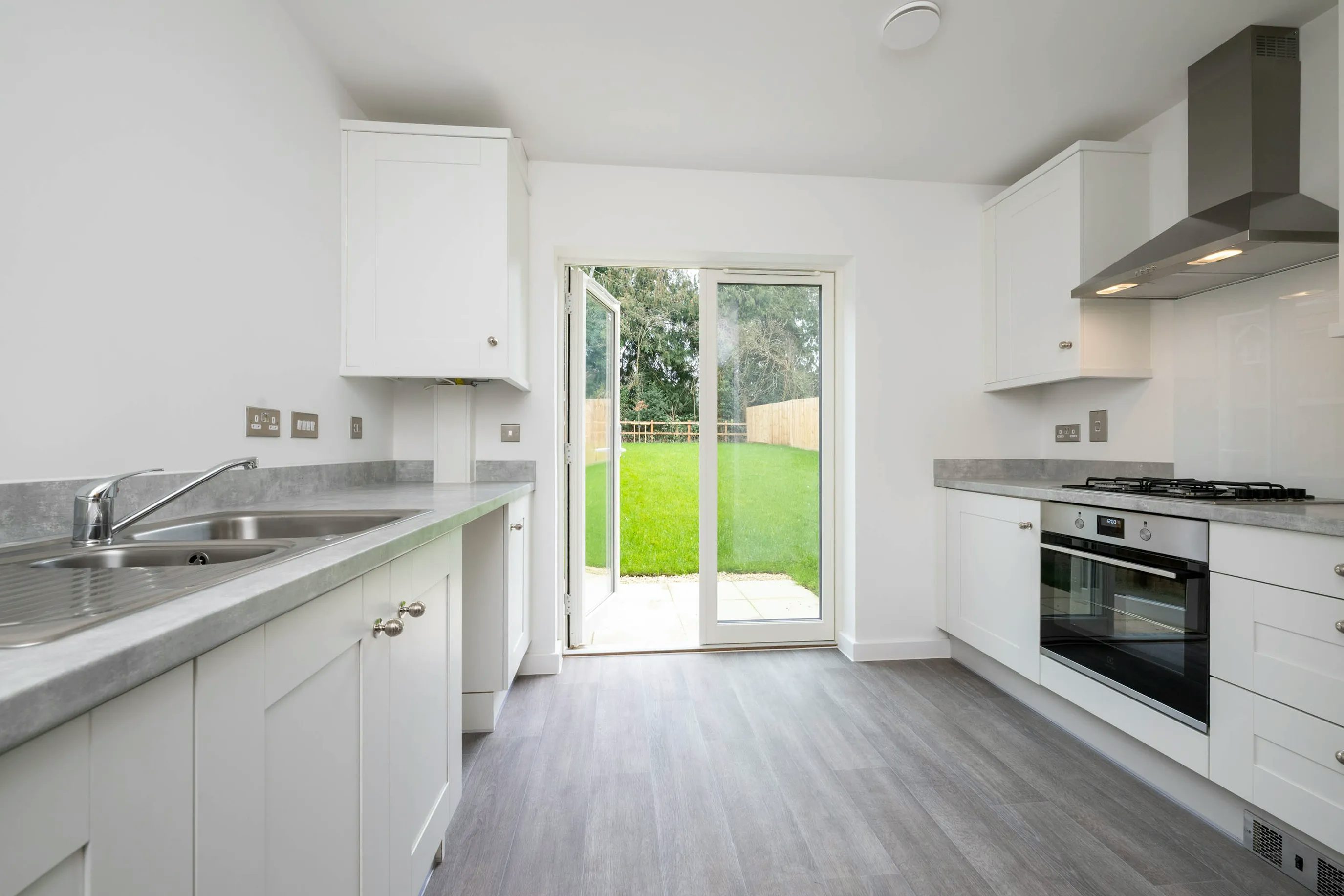 A bright, modern kitchen with white cabinets, grey countertops, and stainless steel appliances. A double sink sits on the left beneath a window, while a gas stove with oven and overhead range hood is positioned on the right. Grey flooring leads to a large glass sliding door at the back, revealing a green lawn and letting in natural light