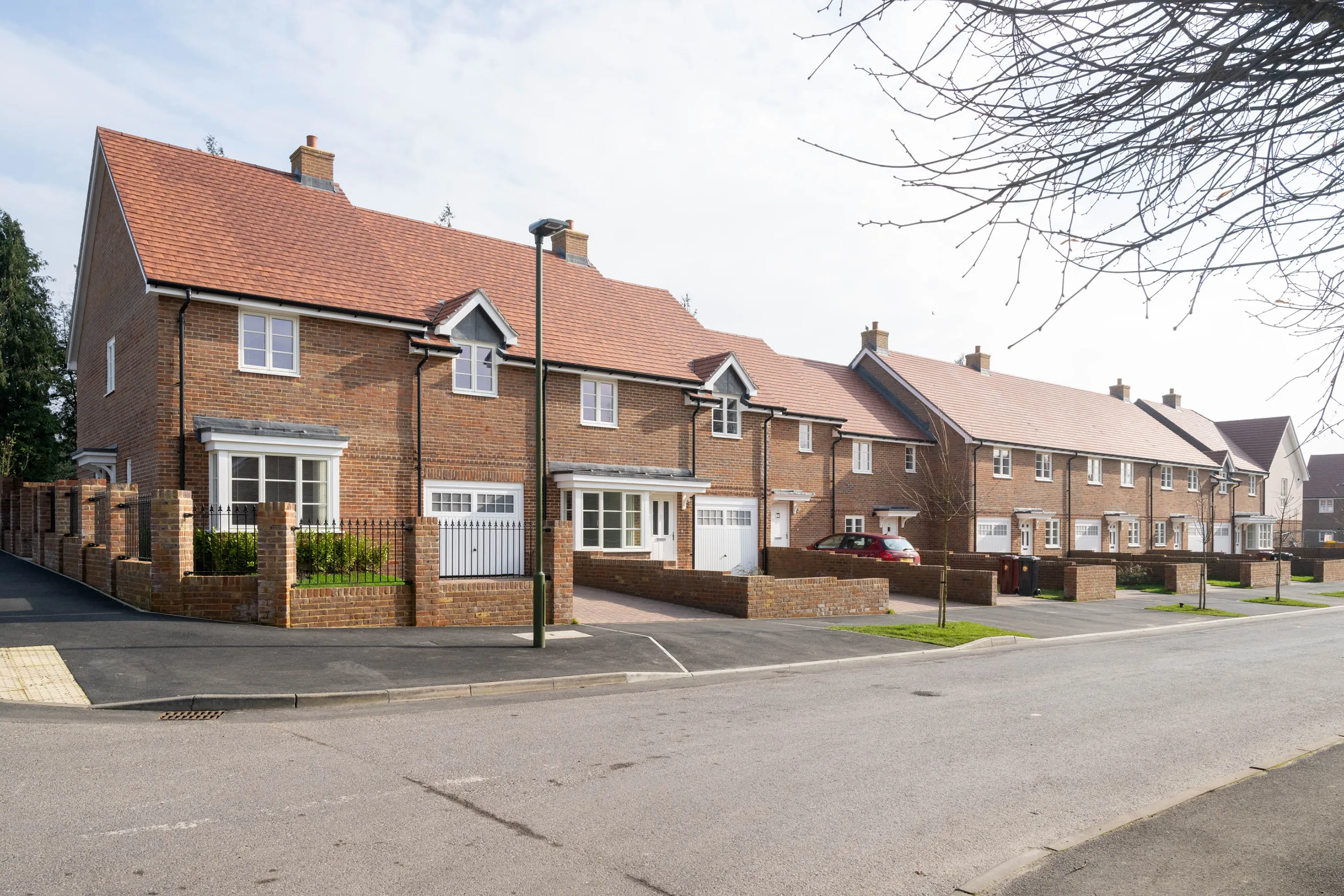 A row of modern red-brick townhouses with red-tiled roofs lines a quiet street, each featuring a small front garden bordered by low brick walls. Several houses have garages, and a few cars are parked along the sidewalk under clear daylight. The scene is residential and neatly maintained.