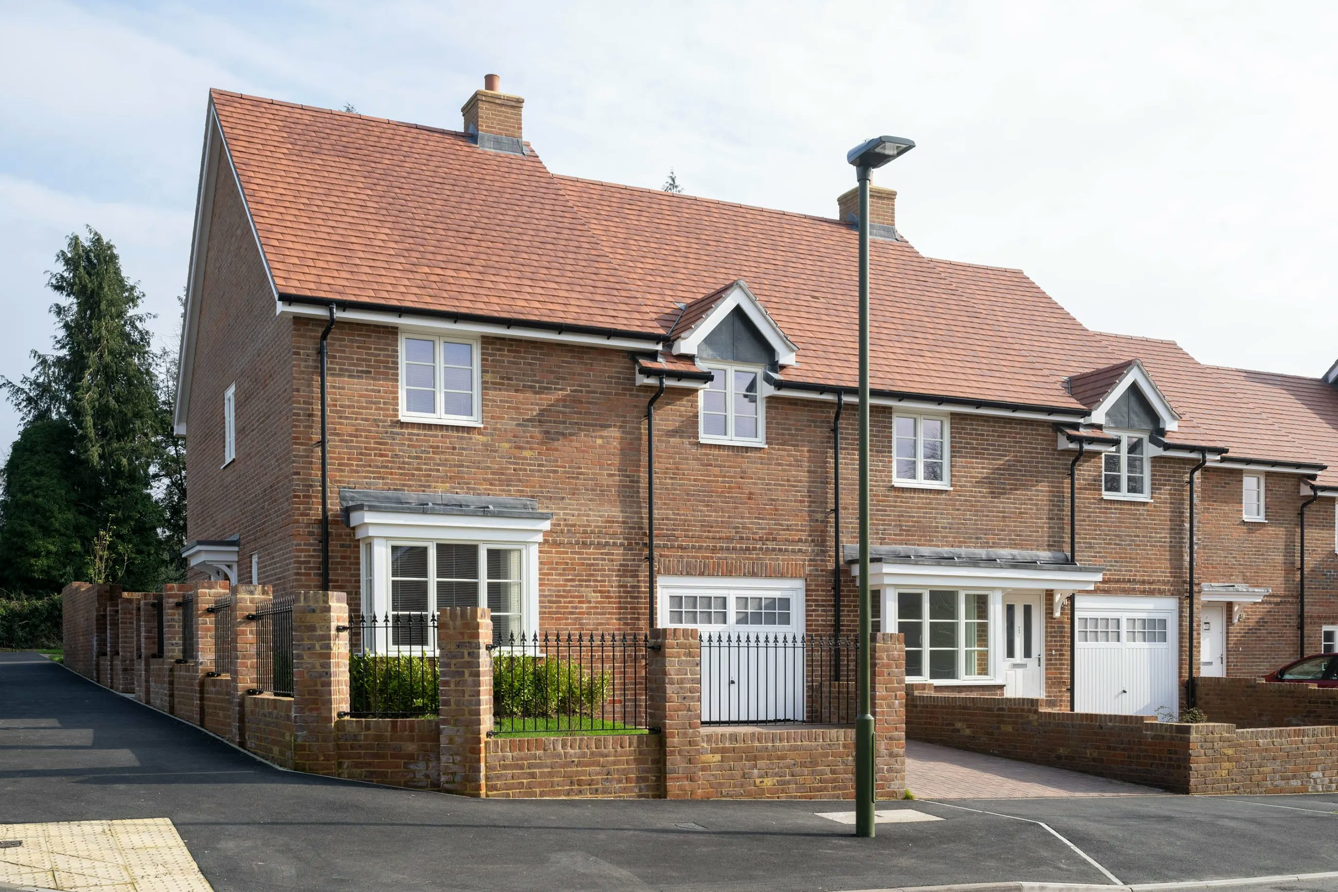 A row of modern two-story brick townhouses with pitched roofs, chimneys, and symmetrical facades. Each home features large front-facing windows, a garage door, and a small garden enclosed by a low brick wall and wrought iron gate. A paved street with a tall streetlamp runs in front, contributing to the neat, residential setting.
