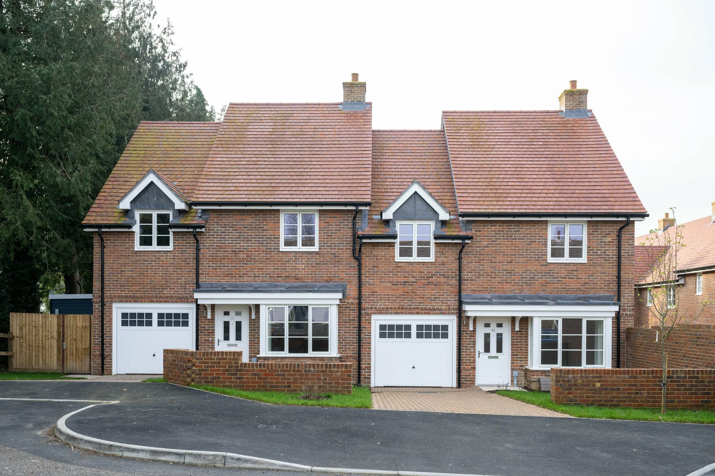 Two identical semi-detached brick houses with pitched tile roofs, each featuring a white garage door, front entrance, and upper and lower windows. The homes share a paved driveway and are set back from the road with small front lawns, in a quiet suburban setting.