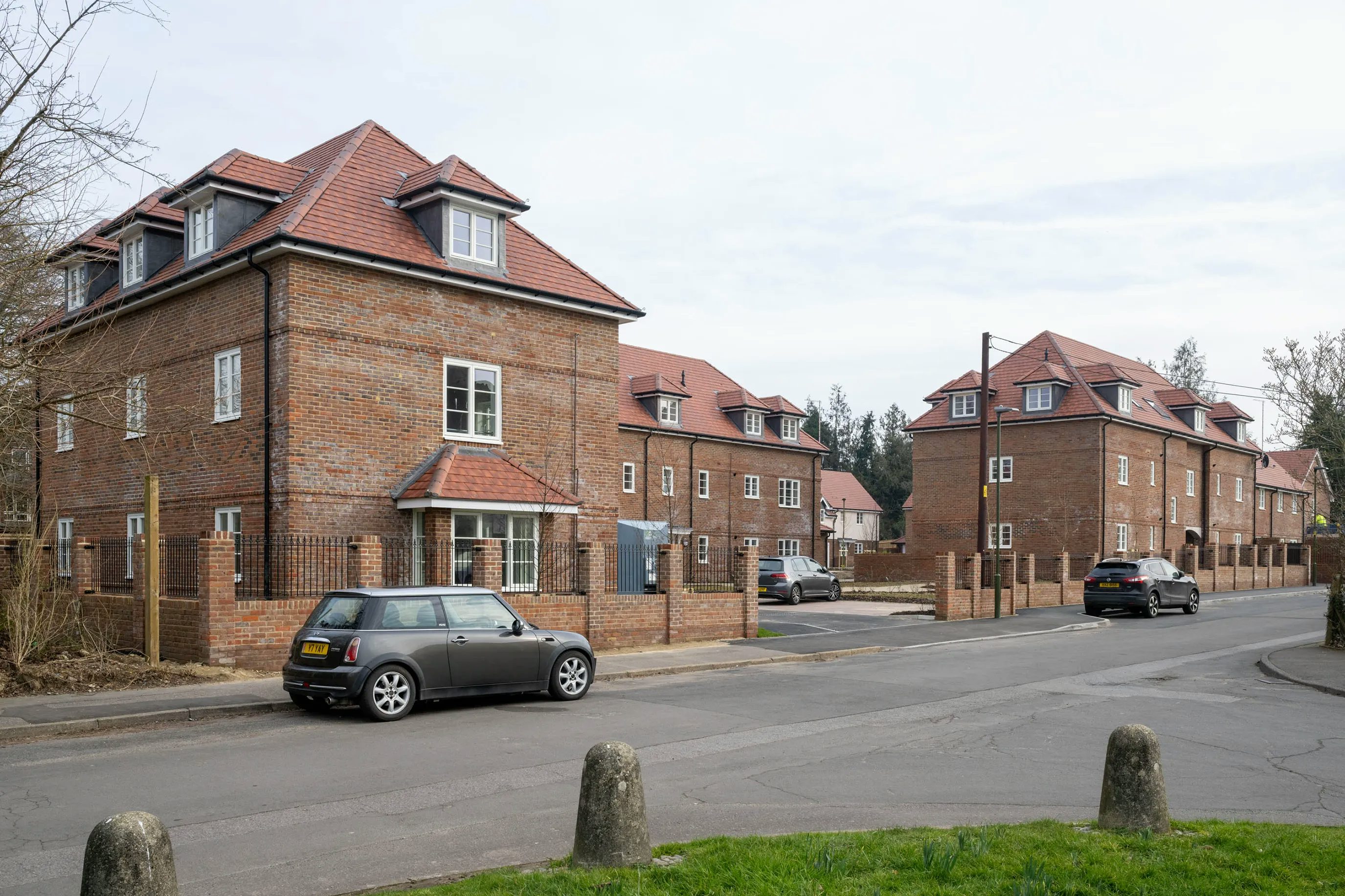 Suburban street view featuring several red-brick houses with red-tiled roofs and dormer windows. Two parked cars, including a Mini Cooper, are visible on the quiet street. A low brick wall and grassy verge with bollards line the foreground.
