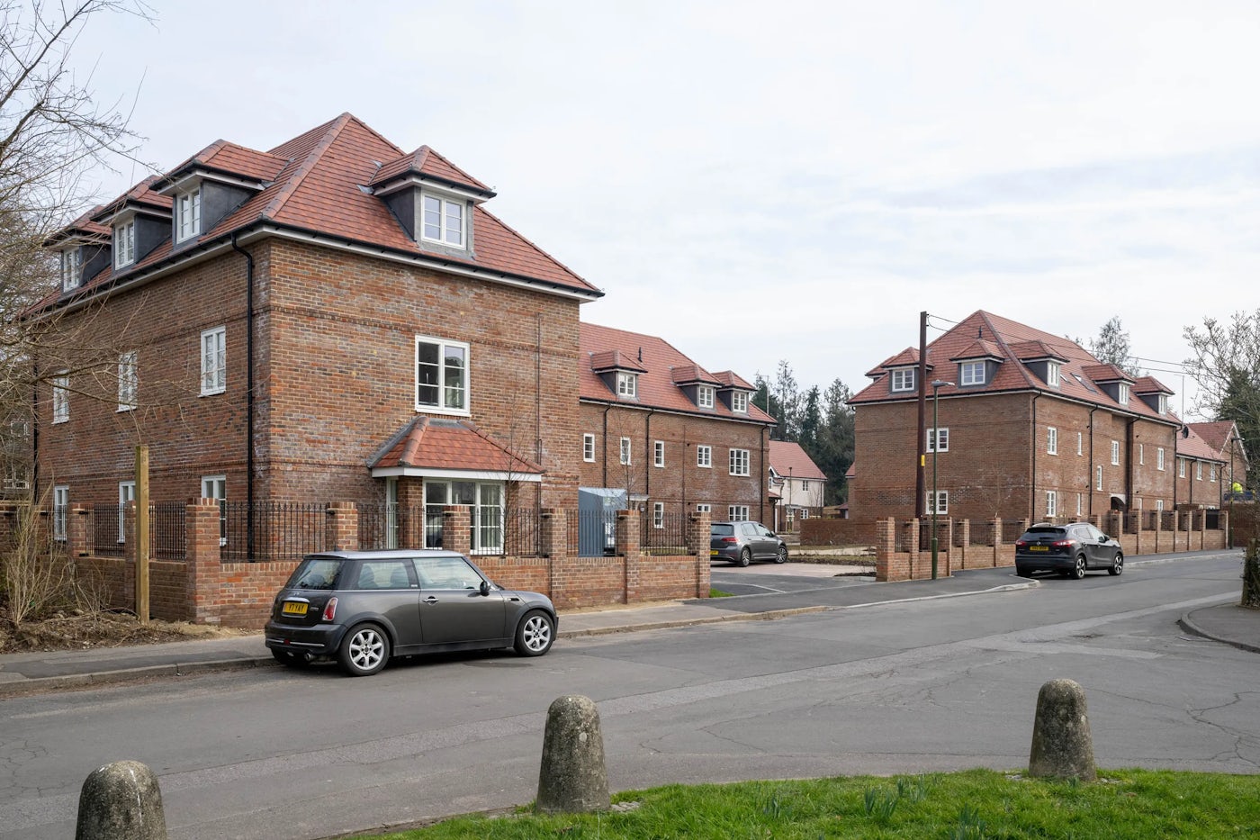 Suburban street view featuring several red-brick houses with red-tiled roofs and dormer windows. Two parked cars, including a Mini Cooper, are visible on the quiet street. A low brick wall and grassy verge with bollards line the foreground.