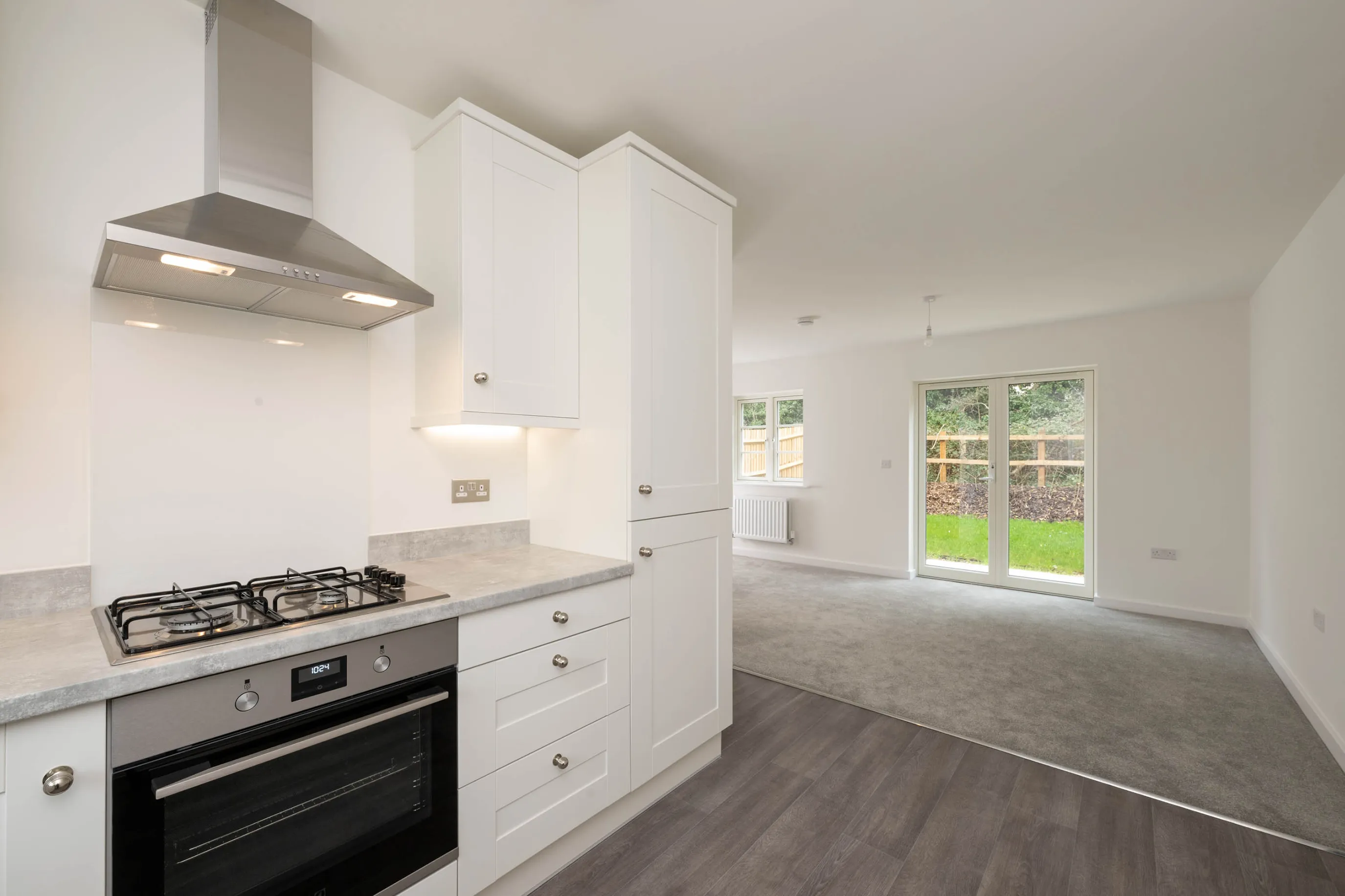 A modern open-plan kitchen and living area with a neutral palette. The kitchen features a stainless steel gas stove, oven, and range hood set against white cabinetry with silver handles and a light grey countertop. Dark wood laminate flooring defines the kitchen zone, while the adjacent living area has grey carpeting. Large windows and a glass door at the back flood the space with natural light and offer views of a garden.