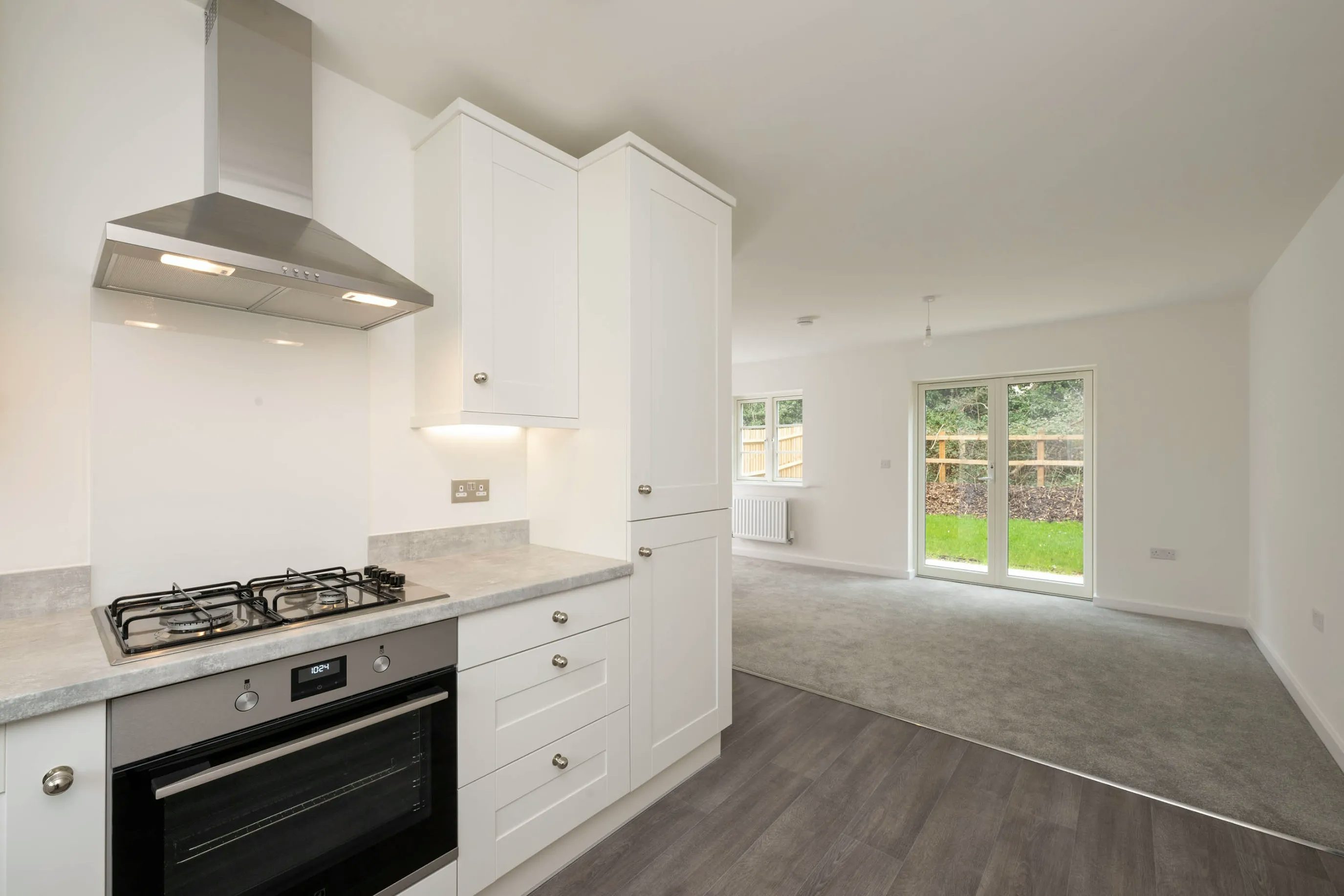 A modern open-plan kitchen and living area with a neutral palette. The kitchen features a stainless steel gas stove, oven, and range hood set against white cabinetry with silver handles and a light grey countertop. Dark wood laminate flooring defines the kitchen zone, while the adjacent living area has grey carpeting. Large windows and a glass door at the back flood the space with natural light and offer views of a garden.