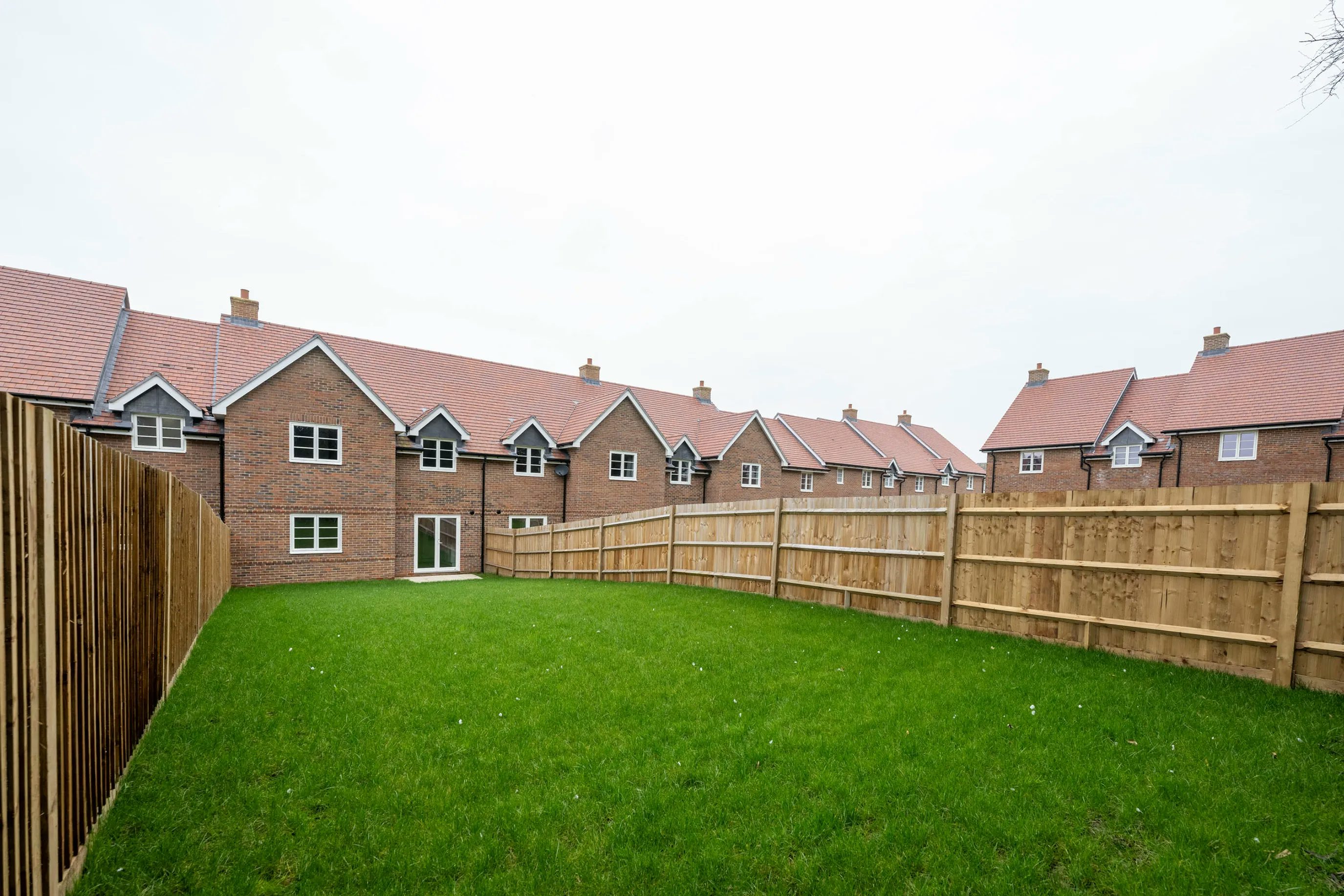 Backyard with a well-kept green lawn enclosed by wooden fences, set against a row of red-brick suburban houses with white-trimmed windows and red-tiled roofs under a cloudy sky.