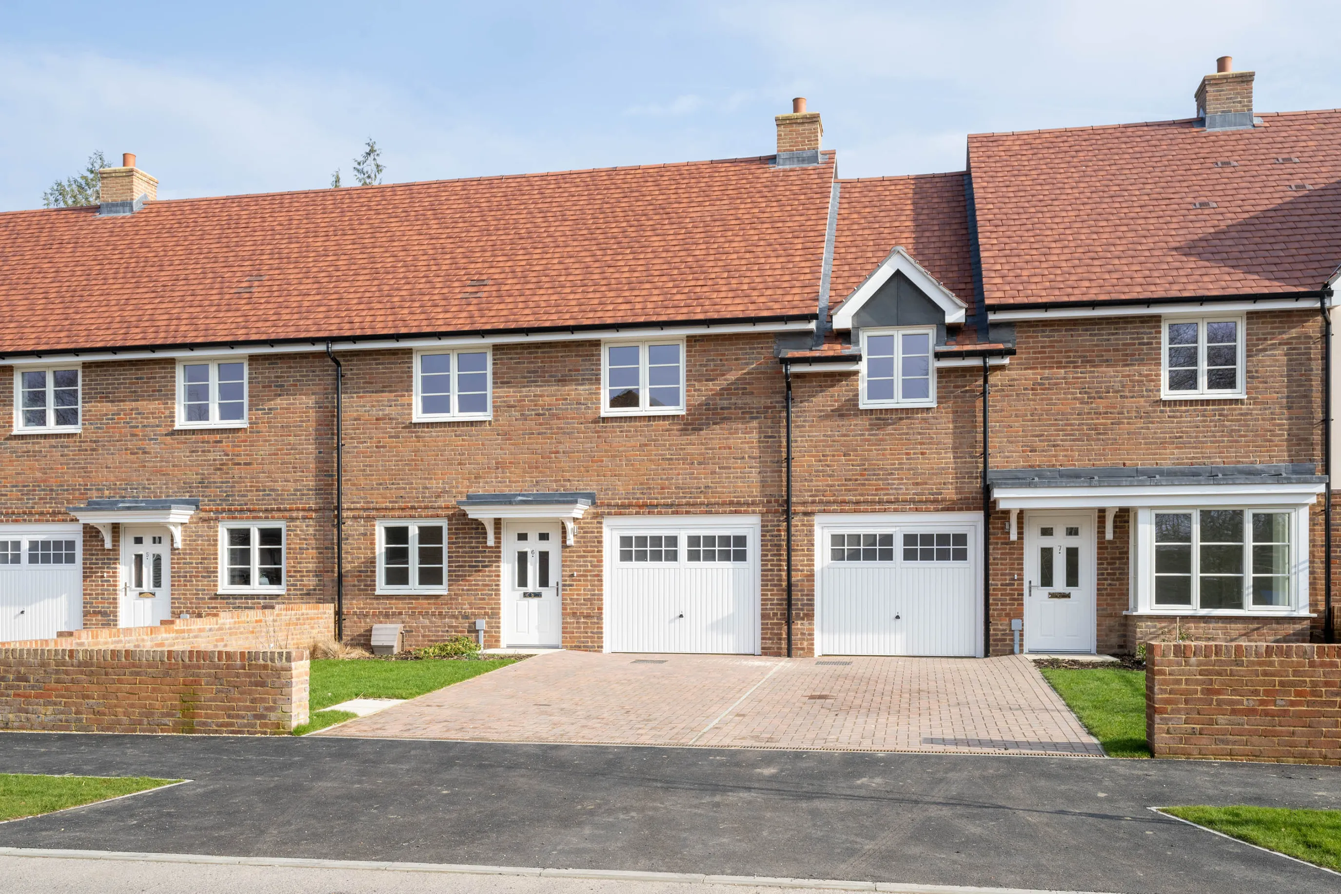 A row of identical two-story townhouses with red-tiled roofs and warm brick exteriors sits beneath a clear blue sky. Each home features a white garage and front door framed by small porches. Paved driveways of light-toned bricks lead up to the houses, flanked by neat patches of grass. Upper-floor windows mirror those on the ground level, creating a balanced, uniform façade across the modern development.