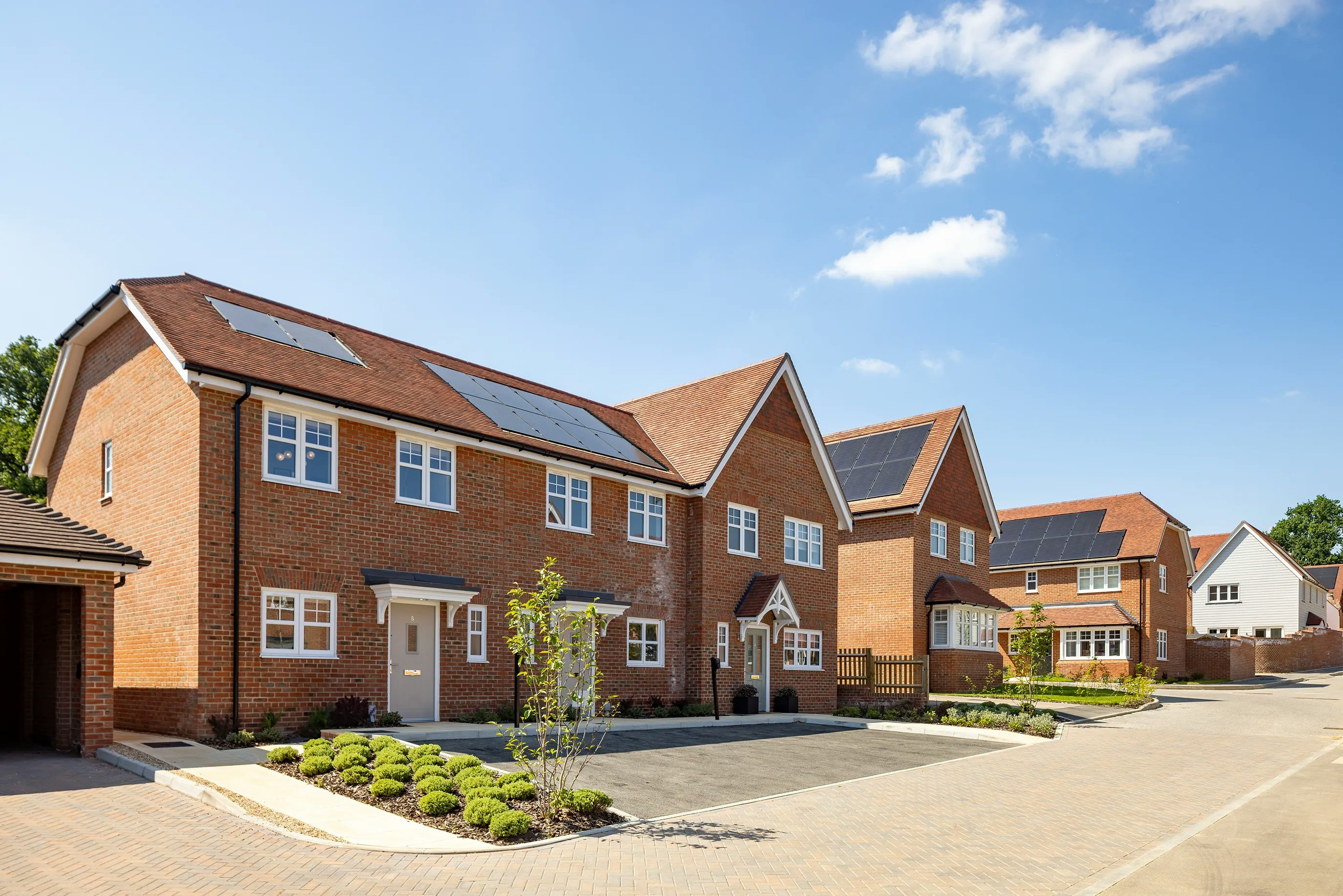 A row of modern brick houses with pitched roofs, each fitted with solar panels, lines a quiet residential street. Manicured front gardens with small shrubs and trees soften the paved driveways. The sky is mostly clear, casting sunlight across the eco-friendly rooftops.