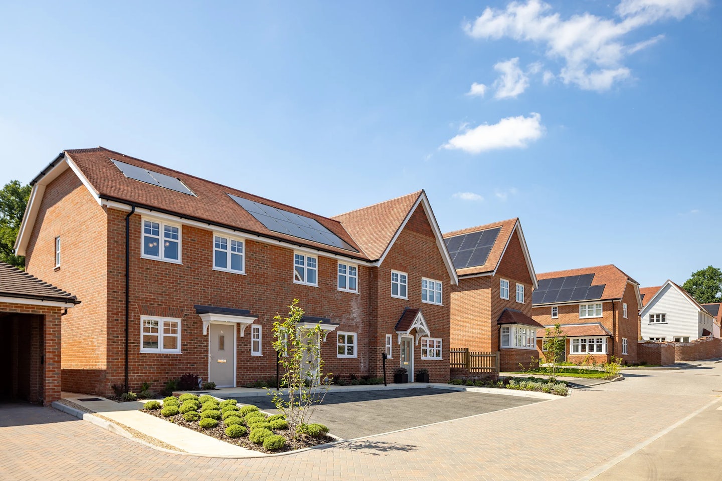 A row of modern brick houses with pitched roofs, each fitted with solar panels, lines a quiet residential street. Manicured front gardens with small shrubs and trees soften the paved driveways. The sky is mostly clear, casting sunlight across the eco-friendly rooftops.