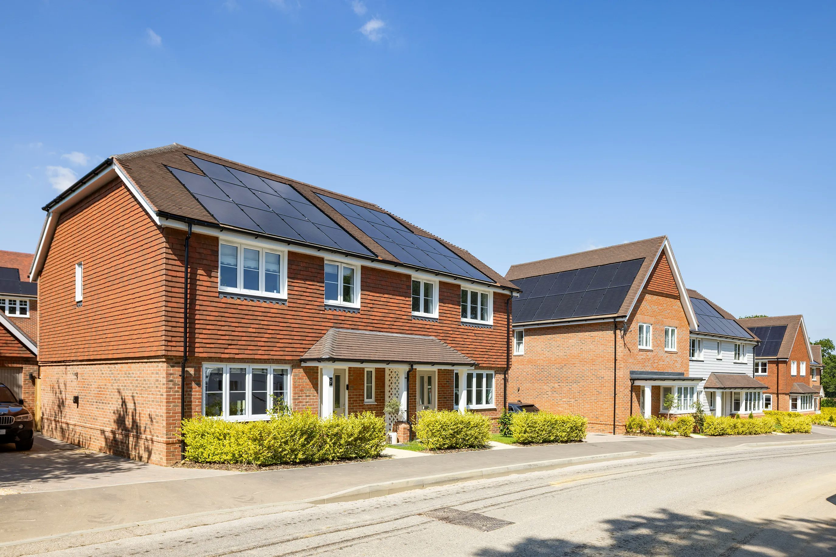 A row of modern, semi-detached brick houses with white-framed windows and doors, each topped with solar panels. The front gardens feature neat hedges and small, well-maintained lawns. Set against a bright blue sky, the scene emphasizes sustainability through renewable energy integration in a suburban neighbourhood.