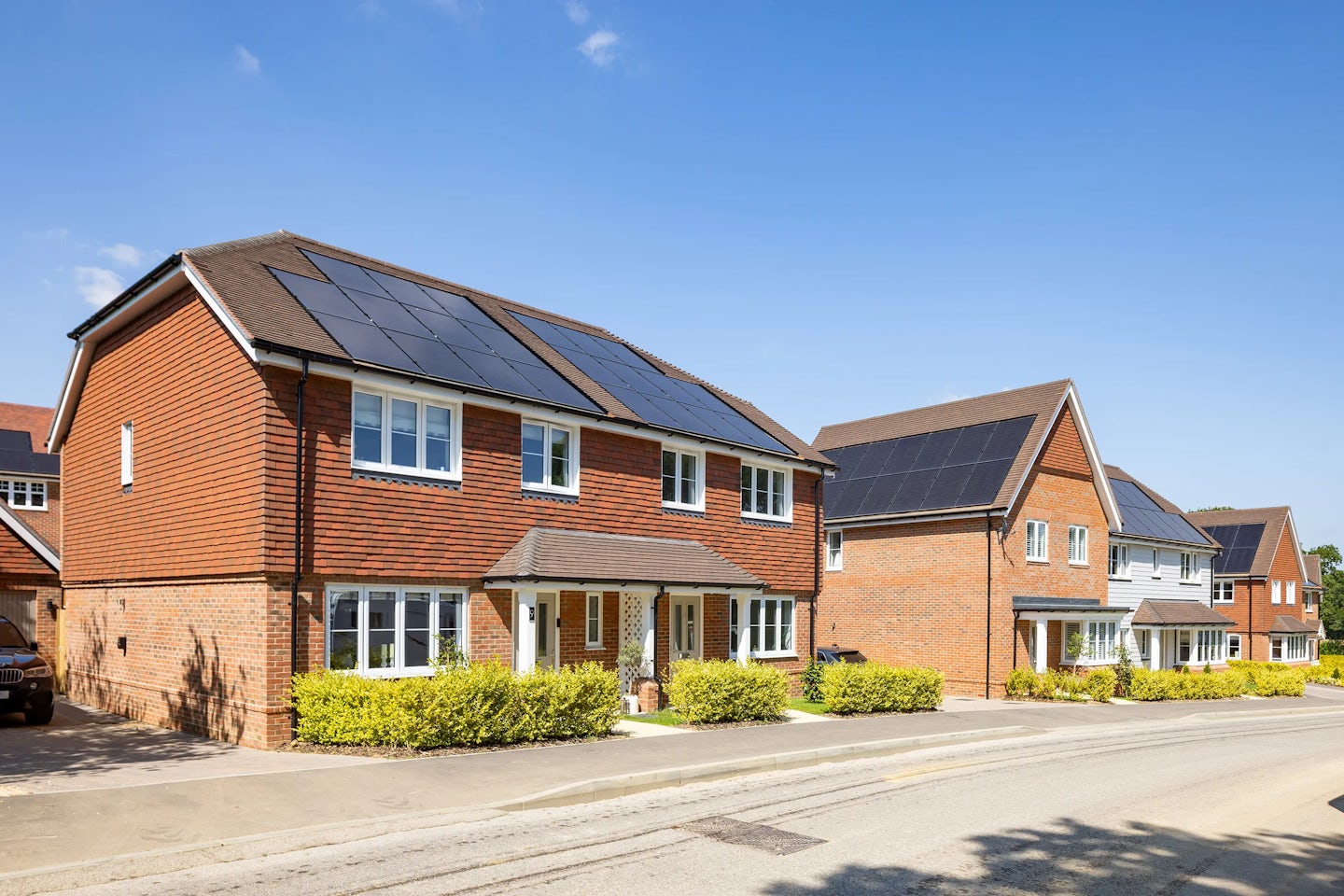 A row of modern, semi-detached brick houses with white-framed windows and doors, each topped with solar panels. The front gardens feature neat hedges and small, well-maintained lawns. Set against a bright blue sky, the scene emphasizes sustainability through renewable energy integration in a suburban neighbourhood.